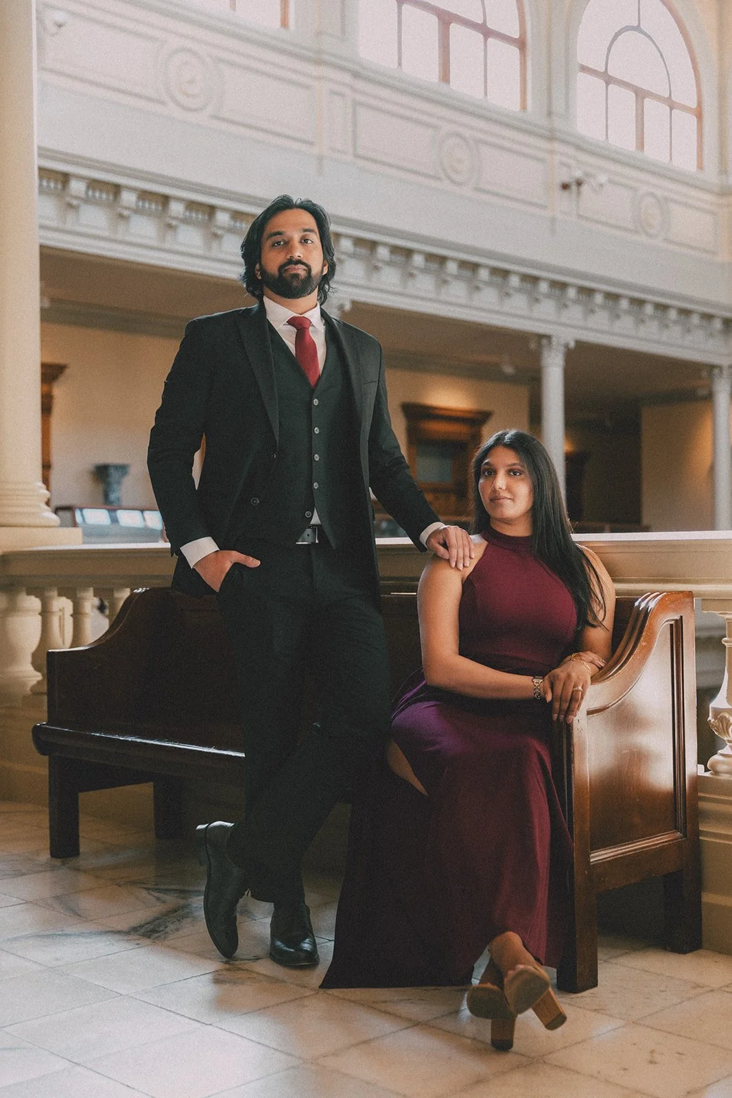 A man in a black suit with a white shirt and red tie standing next to a seated woman in a burgundy dress inside a grand, elegant building with large windows and ornate architecture.