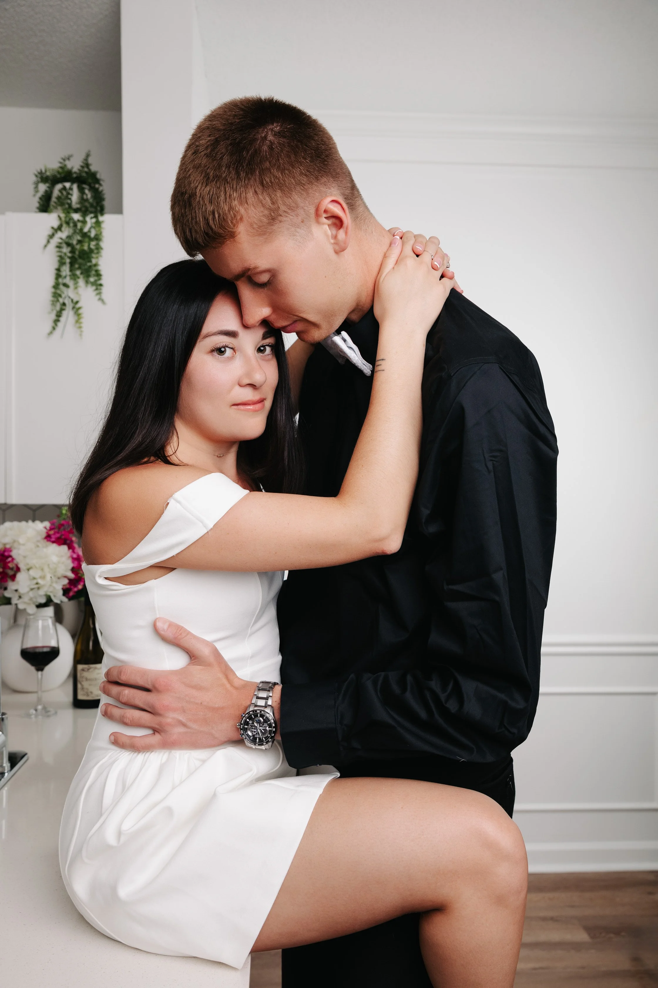 A romantic couple is depicted in an intimate embrace, with the woman sitting on the counter and the man standing, both with eyes closed and foreheads touching, in a bright kitchen with flowers and wine in the background.