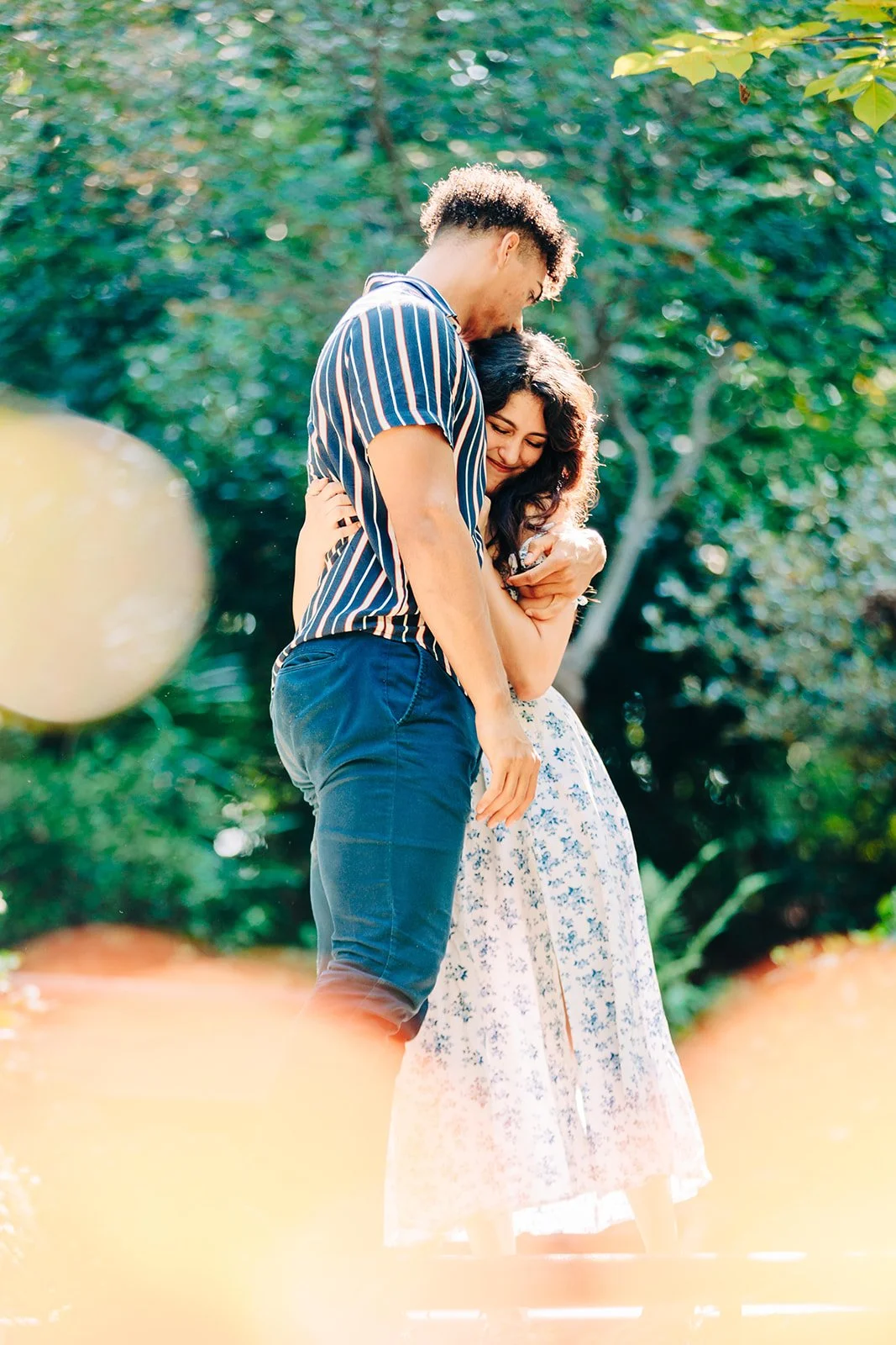 A young couple hugging outdoors in a park with green trees in the background, both smiling and embracing each other.