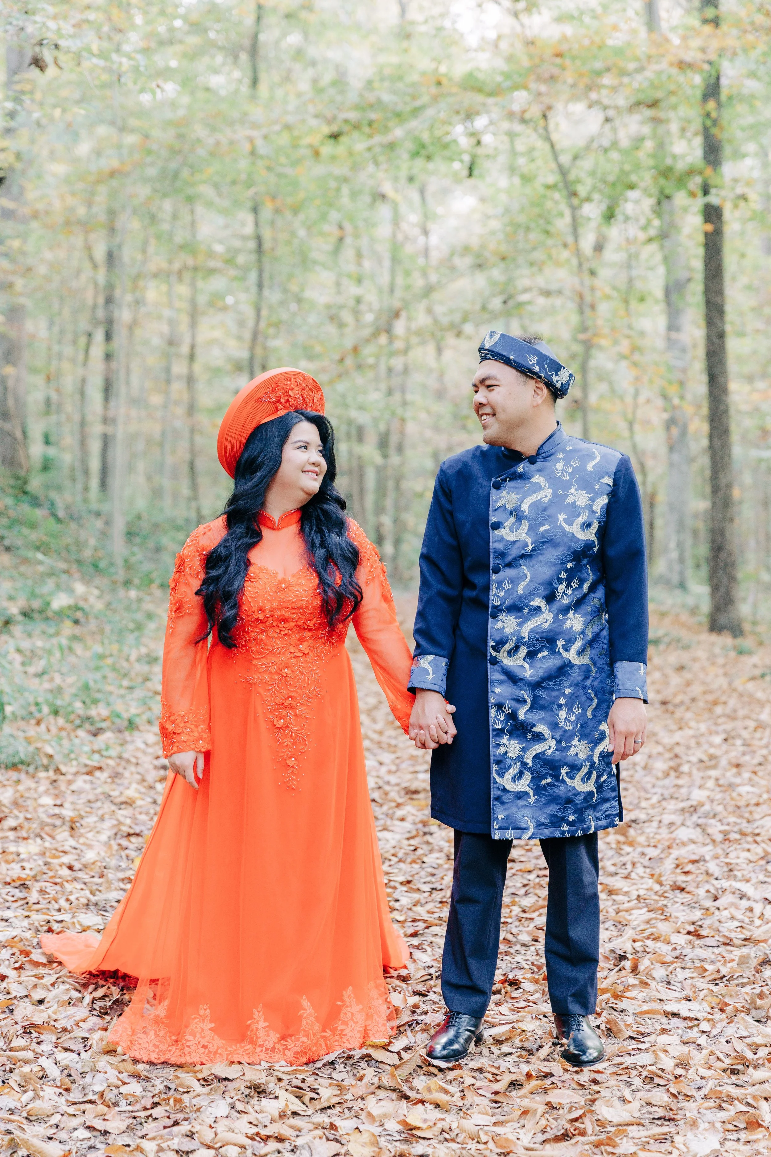 A couple in traditional Vietnamese attire holding hands and smiling in a forest with fallen leaves.