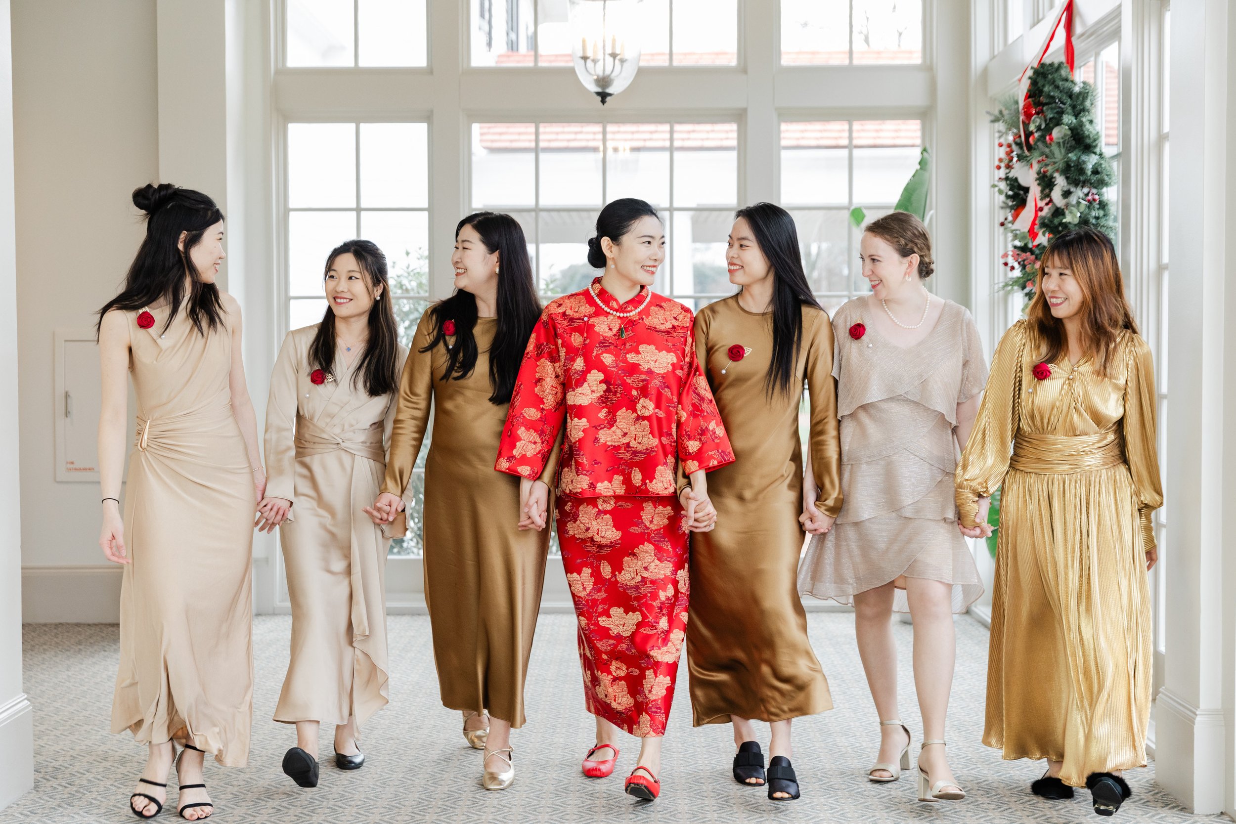 A group of eight women holding hands and walking together indoors, dressed in elegant attire with red roses on their clothing, with a festive Christmas decoration in the background.