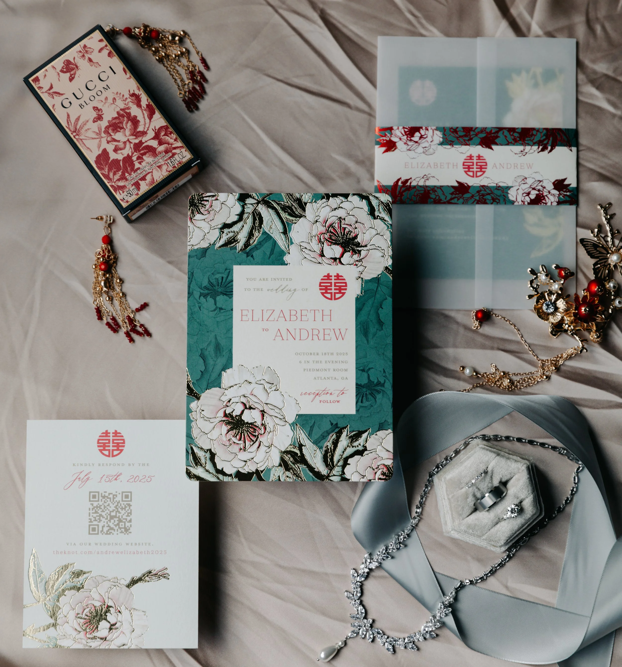 Wedding invitation suite with floral design and gold accents, jewelry including earrings, a necklace, and rings, a silk ribbon, and a Gucci perfume box, all arranged on a beige fabric surface.
