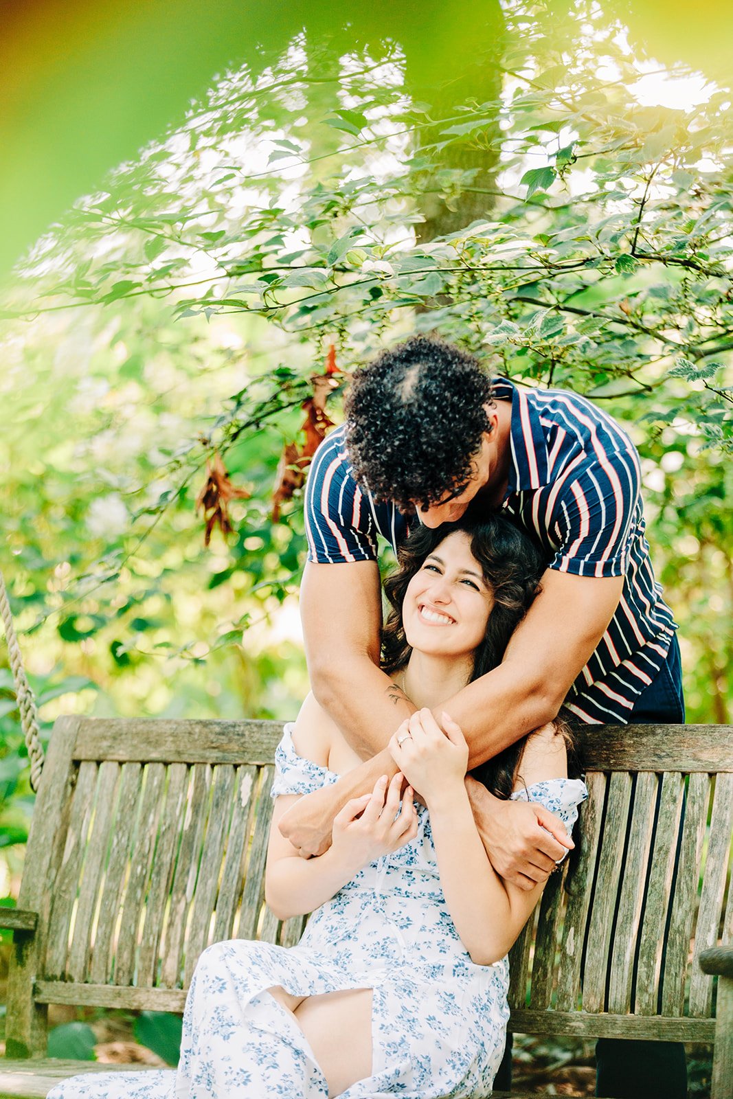 A man and woman sharing a joyful moment on a wooden bench outdoors, surrounded by green foliage.