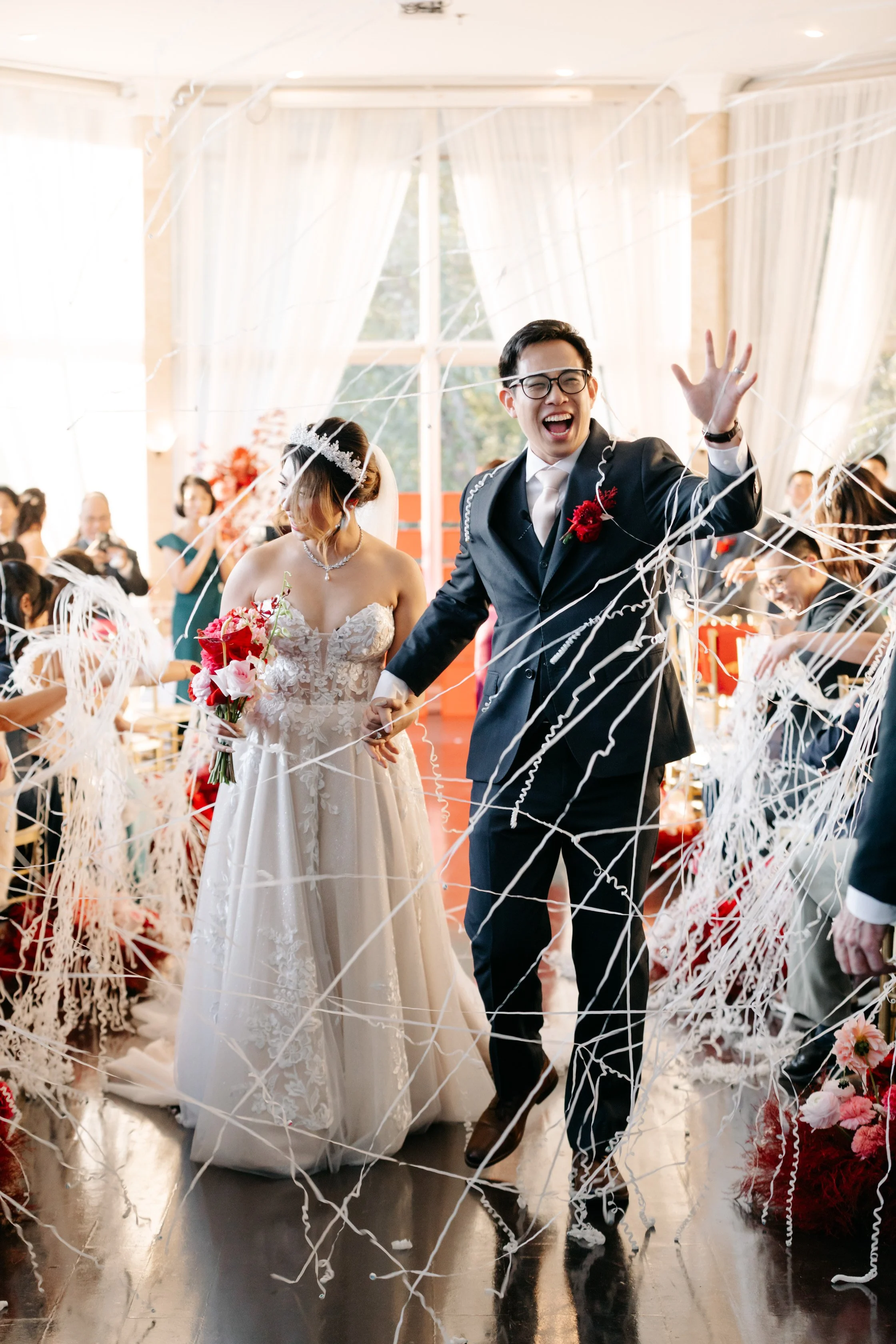 A newlywed couple walking down the aisle during a wedding celebration, surrounded by streamers and guests, with the groom smiling and raising his hand. Fluence Photo & Video. Fluence Wedding.