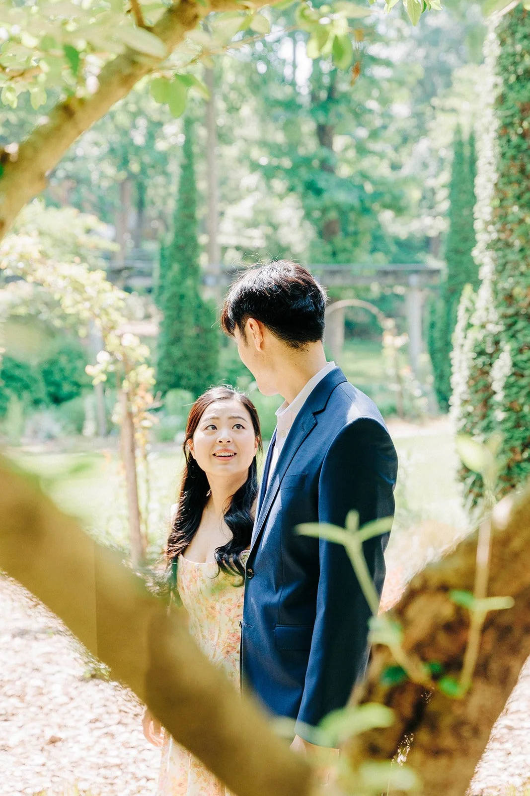 A woman and man standing outdoors in a green, garden-like setting, with trees and plants, engaging in a conversation.