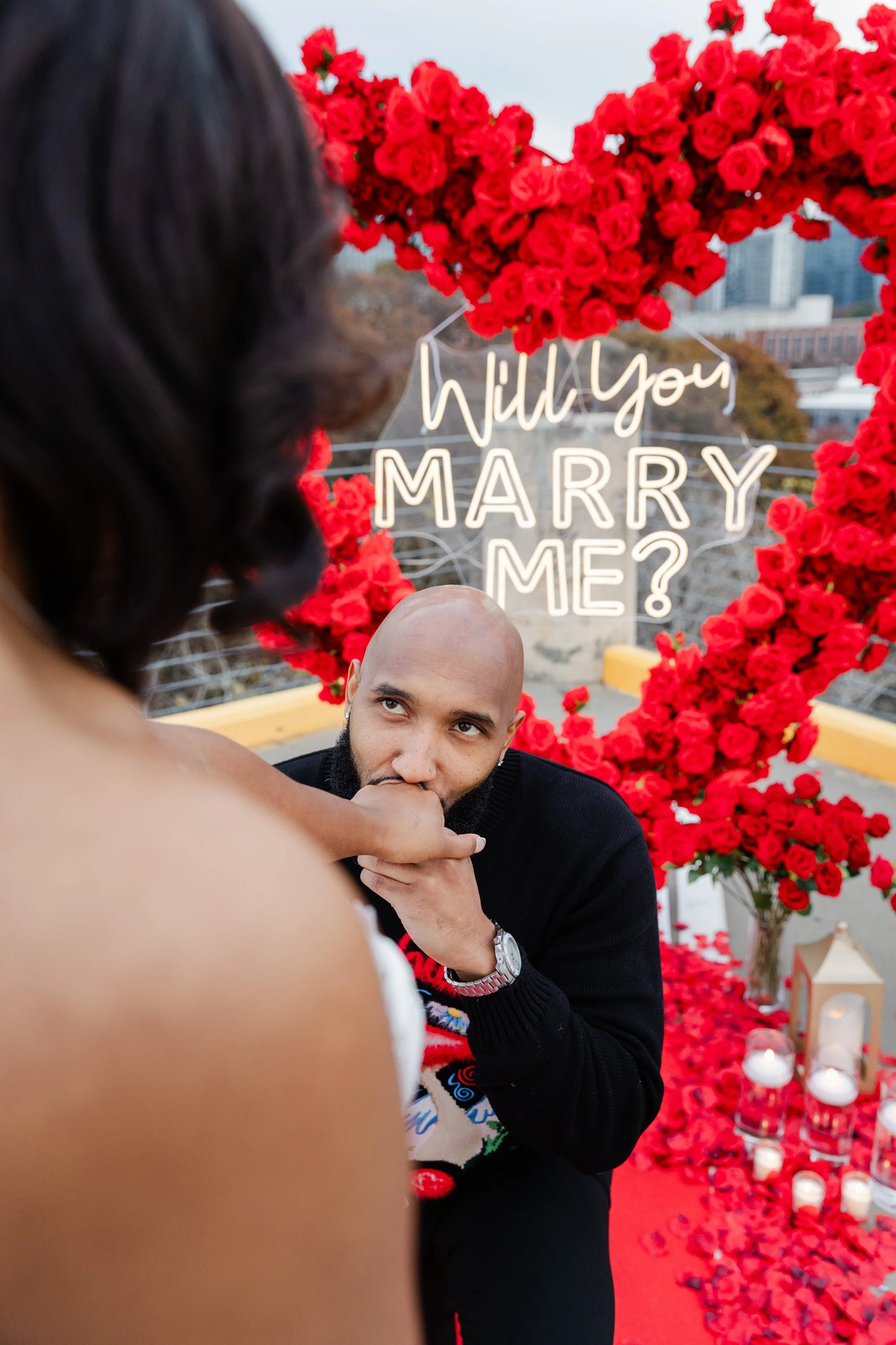A man is kneeling and holding a woman's hand during a proposal on a decorated rooftop with red roses, candles, and a neon sign that reads 'Will you MARRY ME?' in the background.