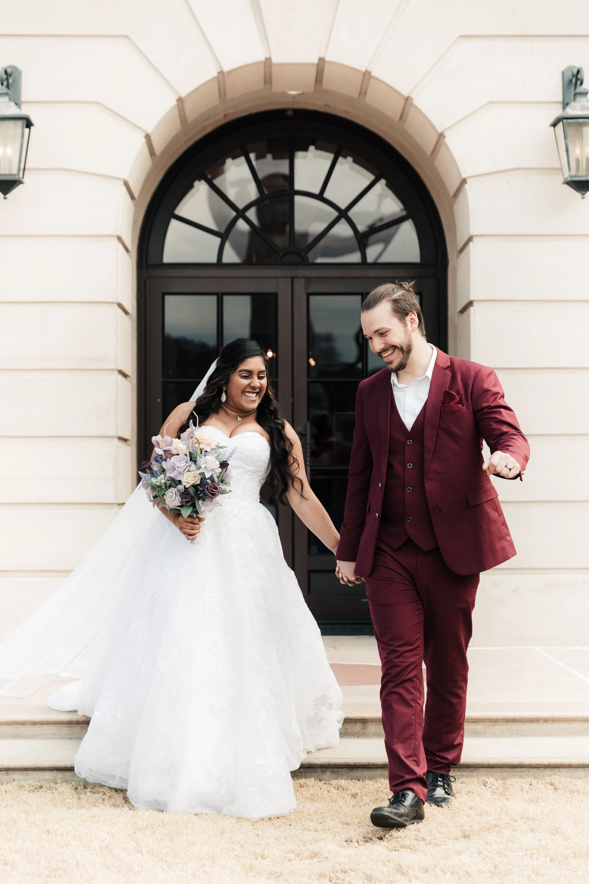 A newlywed couple holding hands, smiling and walking in front of a building with an arched window, with the bride in a white wedding dress holding a bouquet and the groom in a burgundy suit.