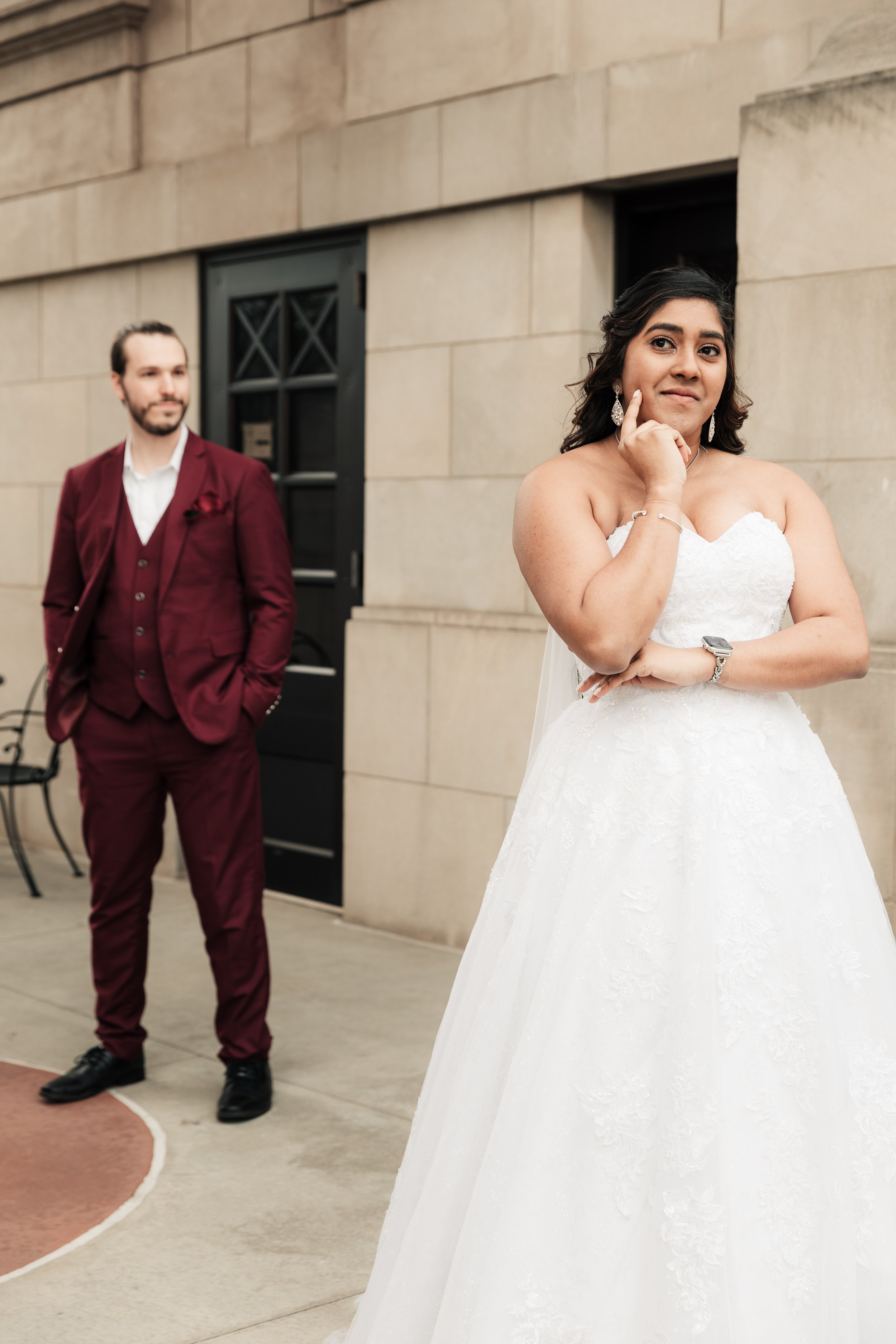 A bride in a white wedding dress with earrings and a bracelet stands with her hand on her chin, looking thoughtful. A groom in a burgundy suit with a white shirt and a matching pocket square stands in the background, looking at her. They are outside 