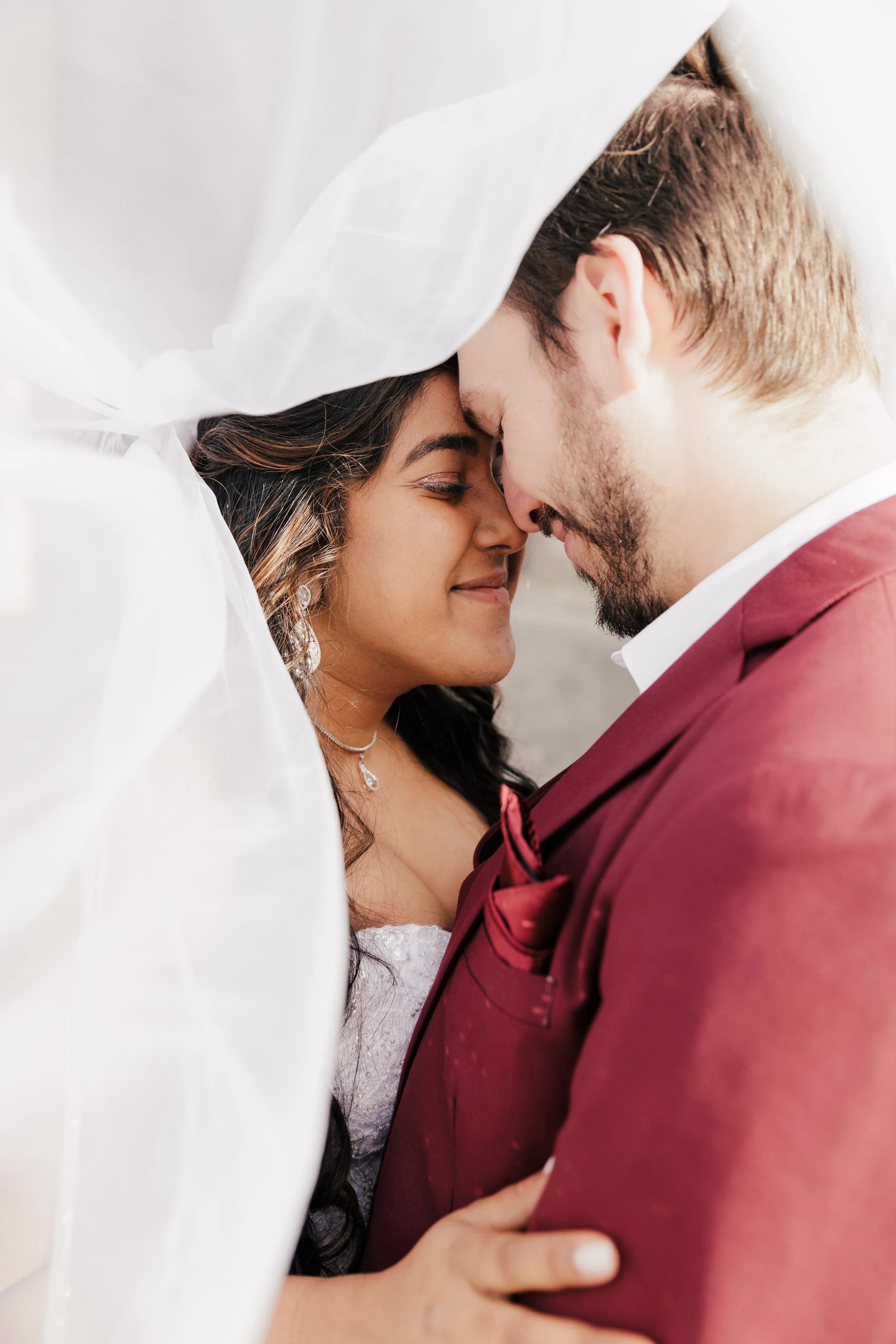 A newlywed couple, a woman with dark hair and a man with light brown hair and beard, share a close moment with their foreheads touching, surrounded by a white veil or fabric during their wedding.