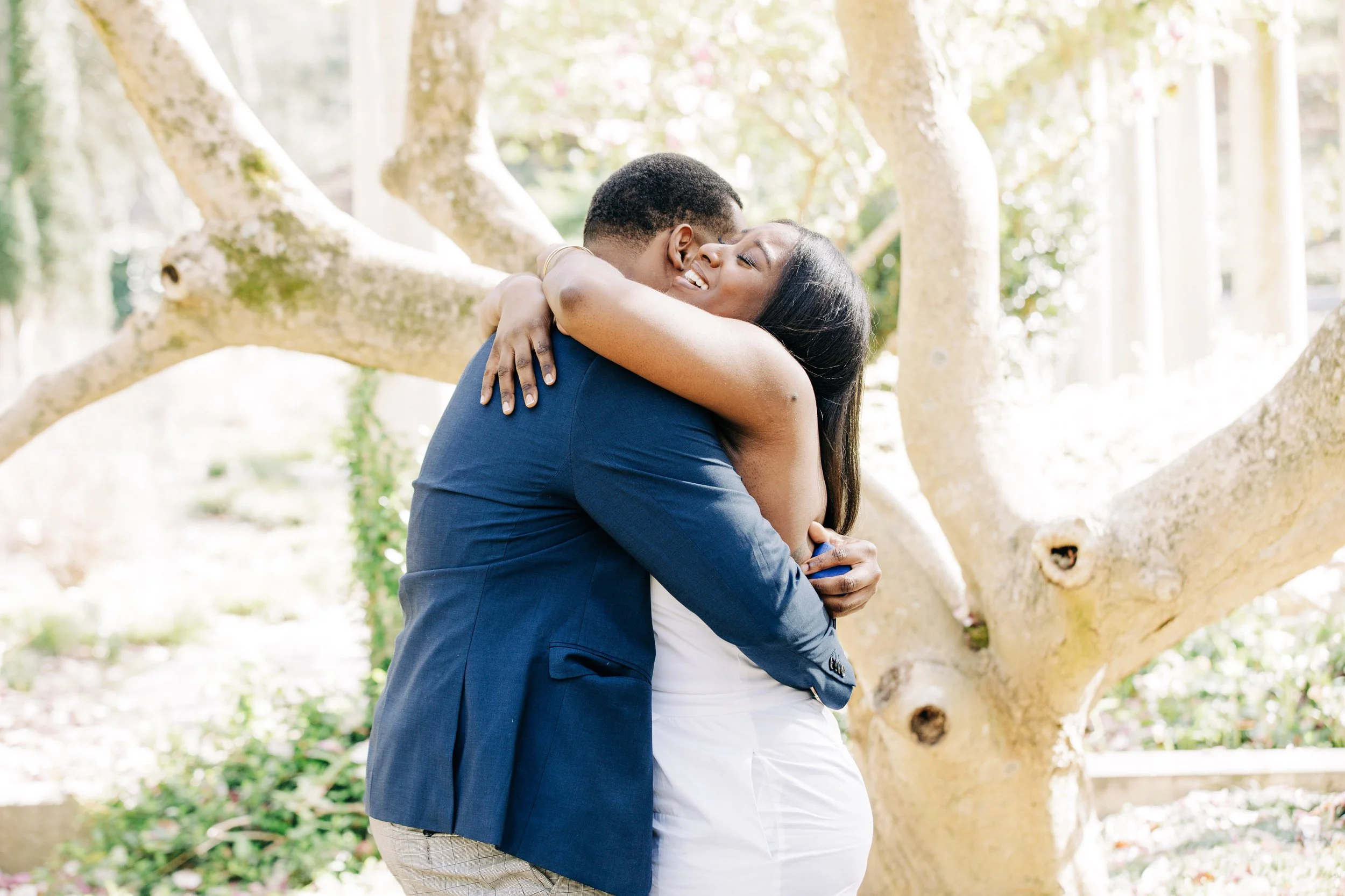 A couple hugging outdoors near a tree, with sunlight filtering through the branches.