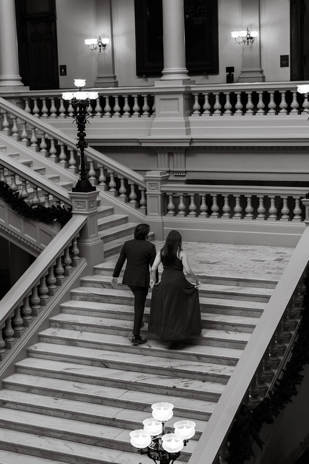 A man and woman in formal attire walking up a grand, marble staircase in a historic building with ornate architectural details and chandeliers.