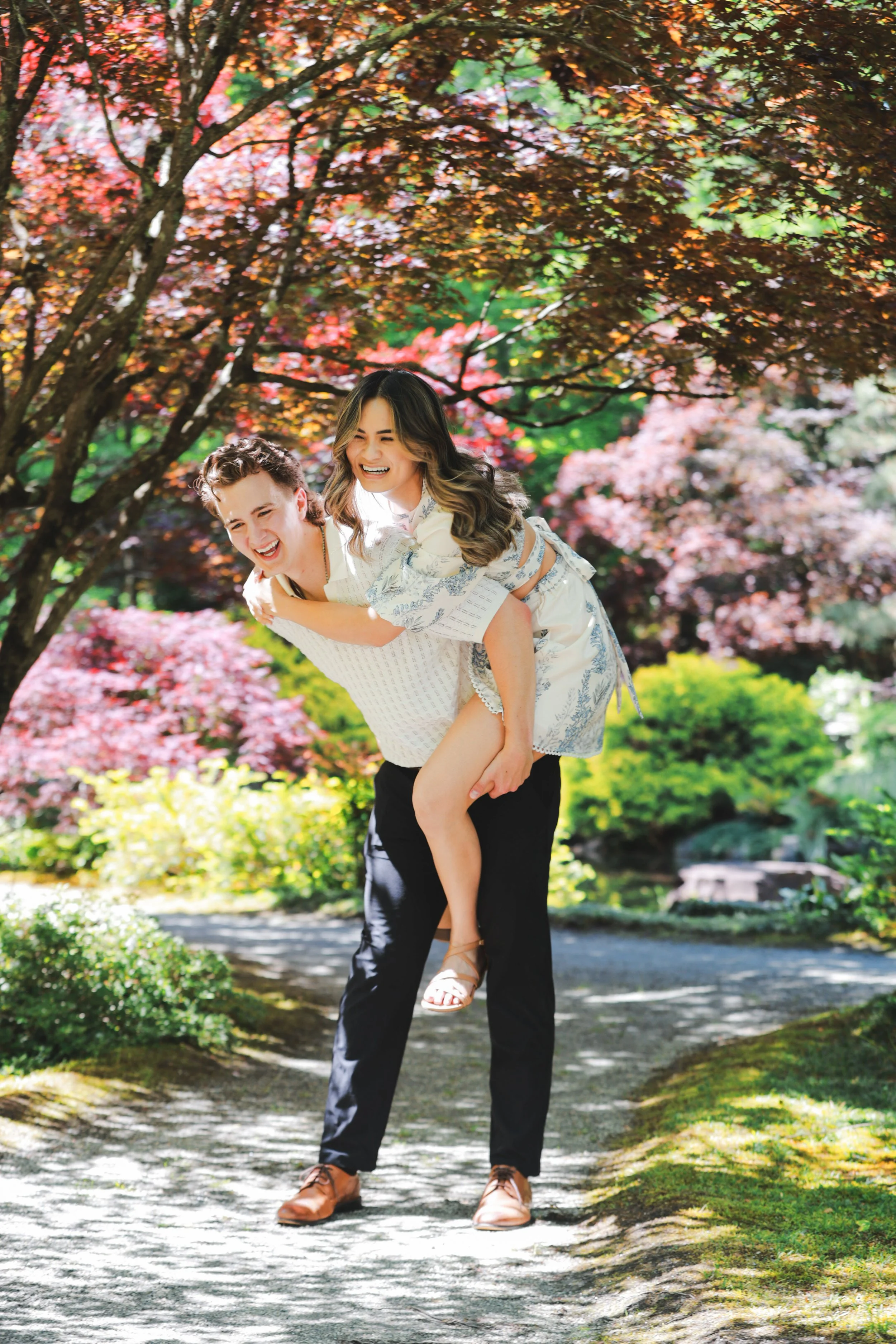 A man giving a woman a piggyback ride on a garden path, surrounded by colorful autumn trees and lush greenery.