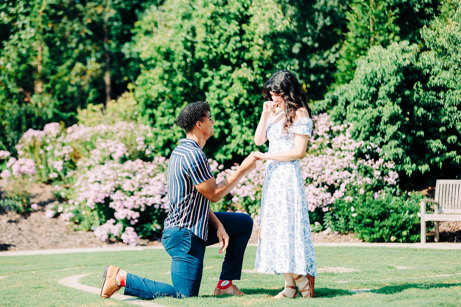 A man proposing marriage to a woman in a garden, with a man kneeling on one knee holding her hand as she reacts happily.