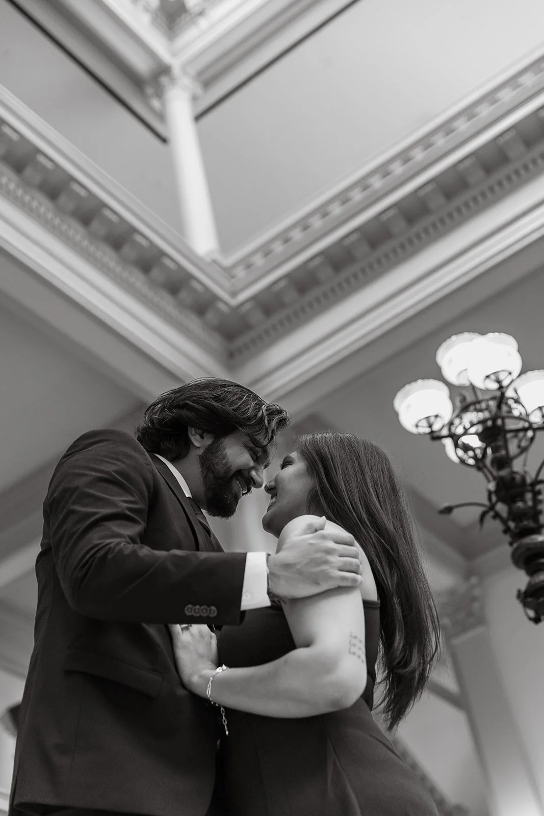 A couple dancing closely indoors, smiling and looking at each other, with ornate ceiling details and a chandelier in the background, in black and white.