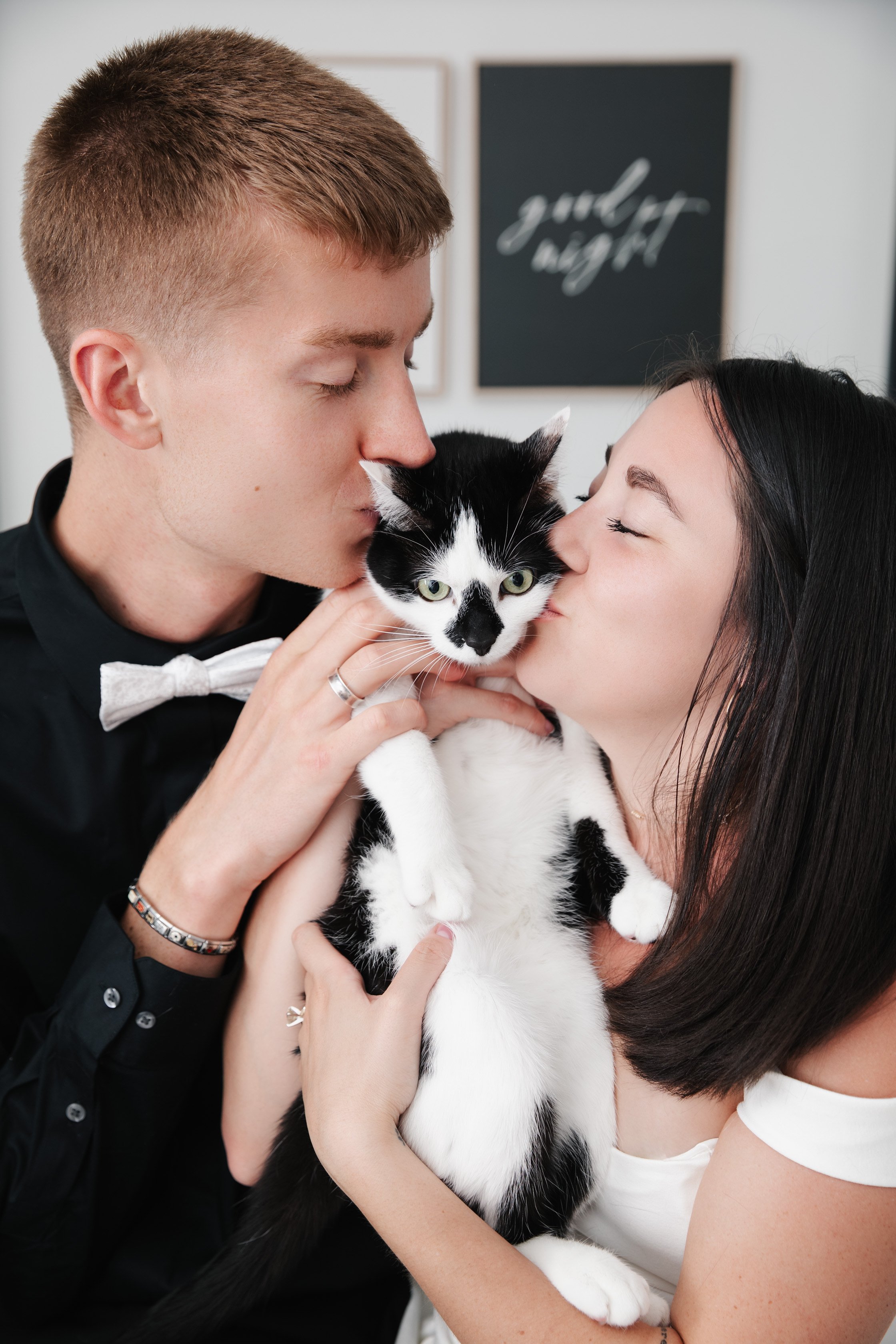 A couple kisses a black and white cat they are holding together.