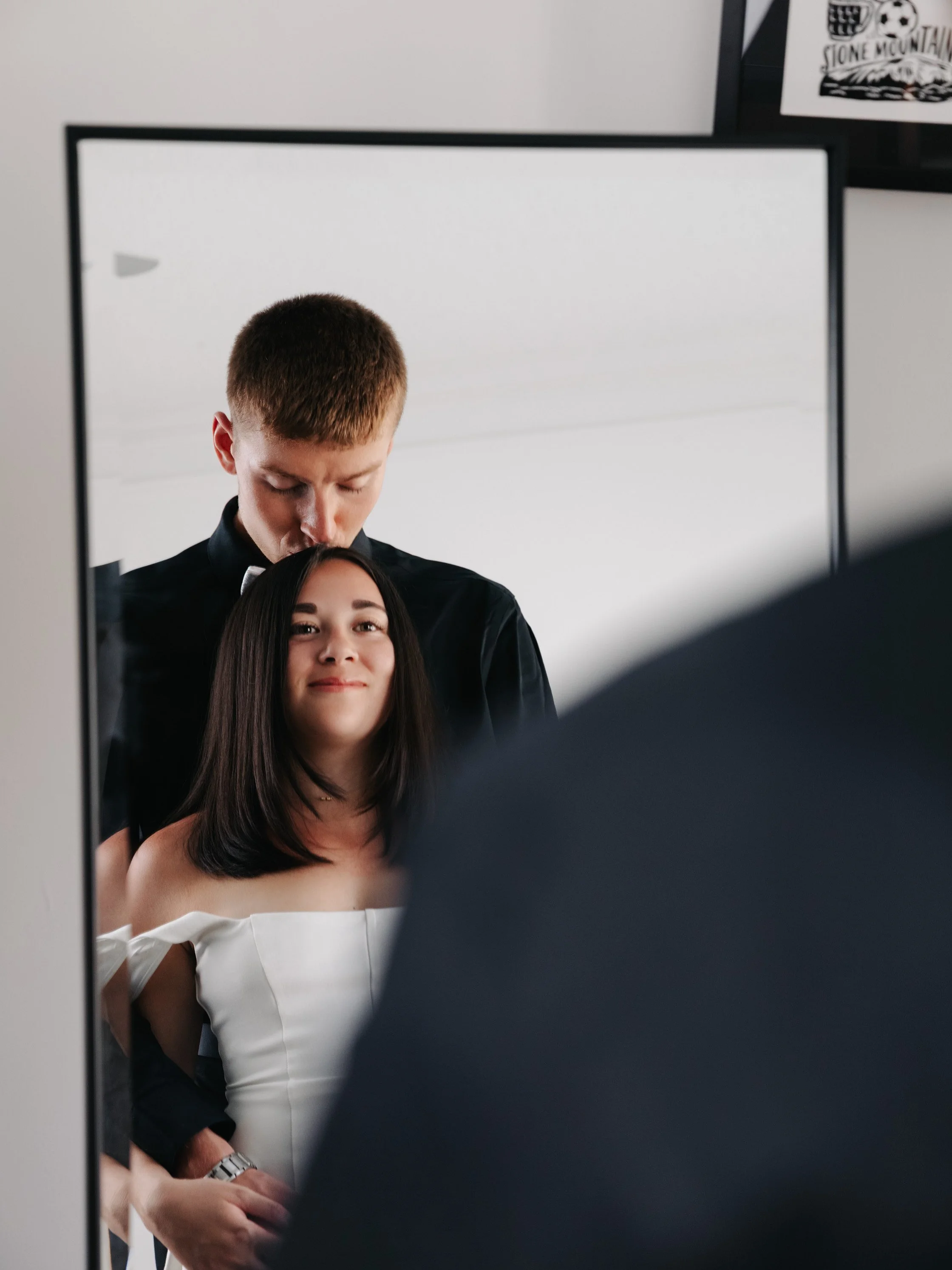 A woman with shoulder-length dark hair and wearing an off-shoulder white dress gets her hair done by a man with short red hair, dressed in black, reflected in a mirror.