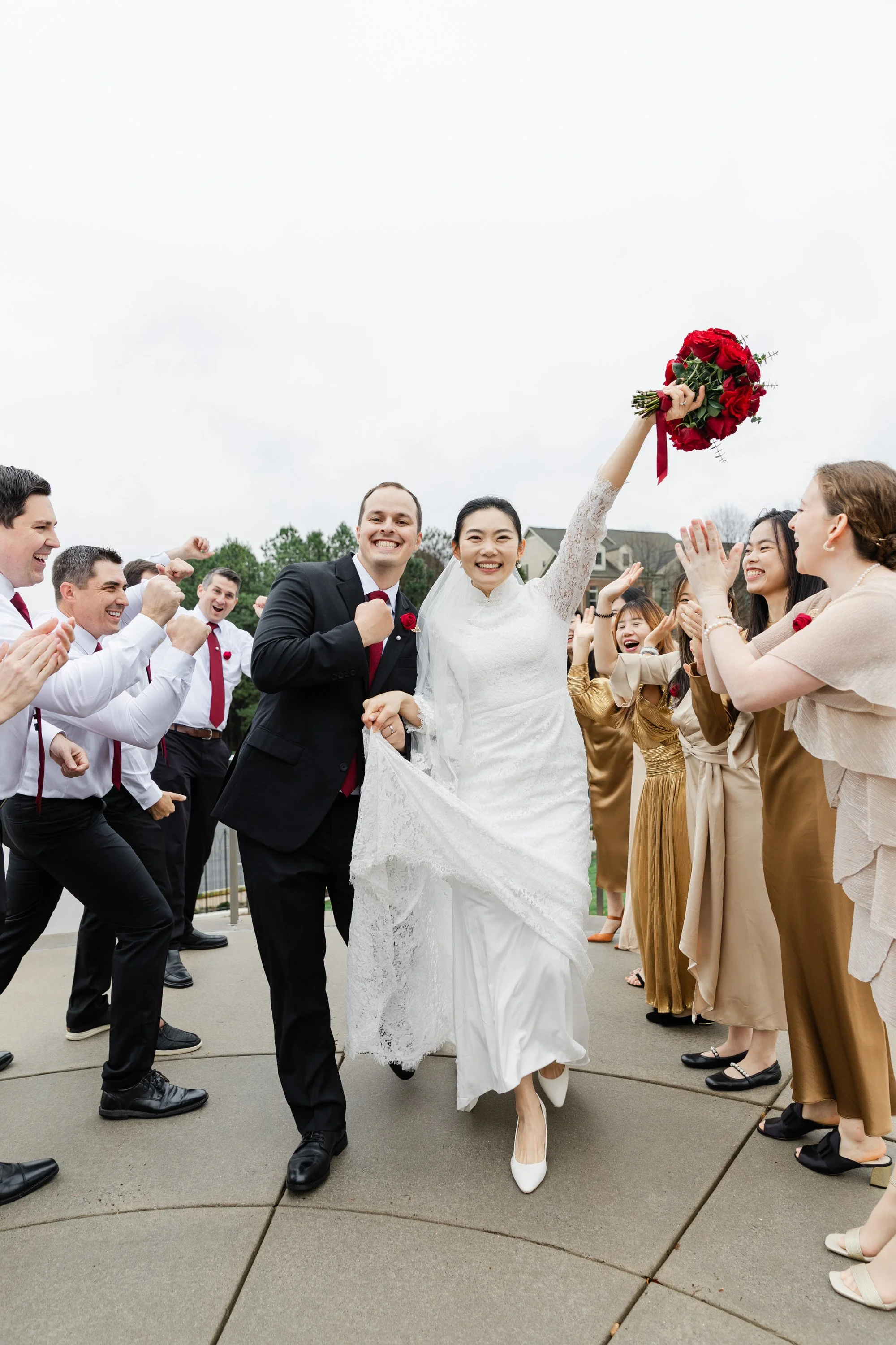 Bride holding a bouquet of red roses with her right hand raised, walking with groom, amid cheering guests outdoors on a cloudy day.