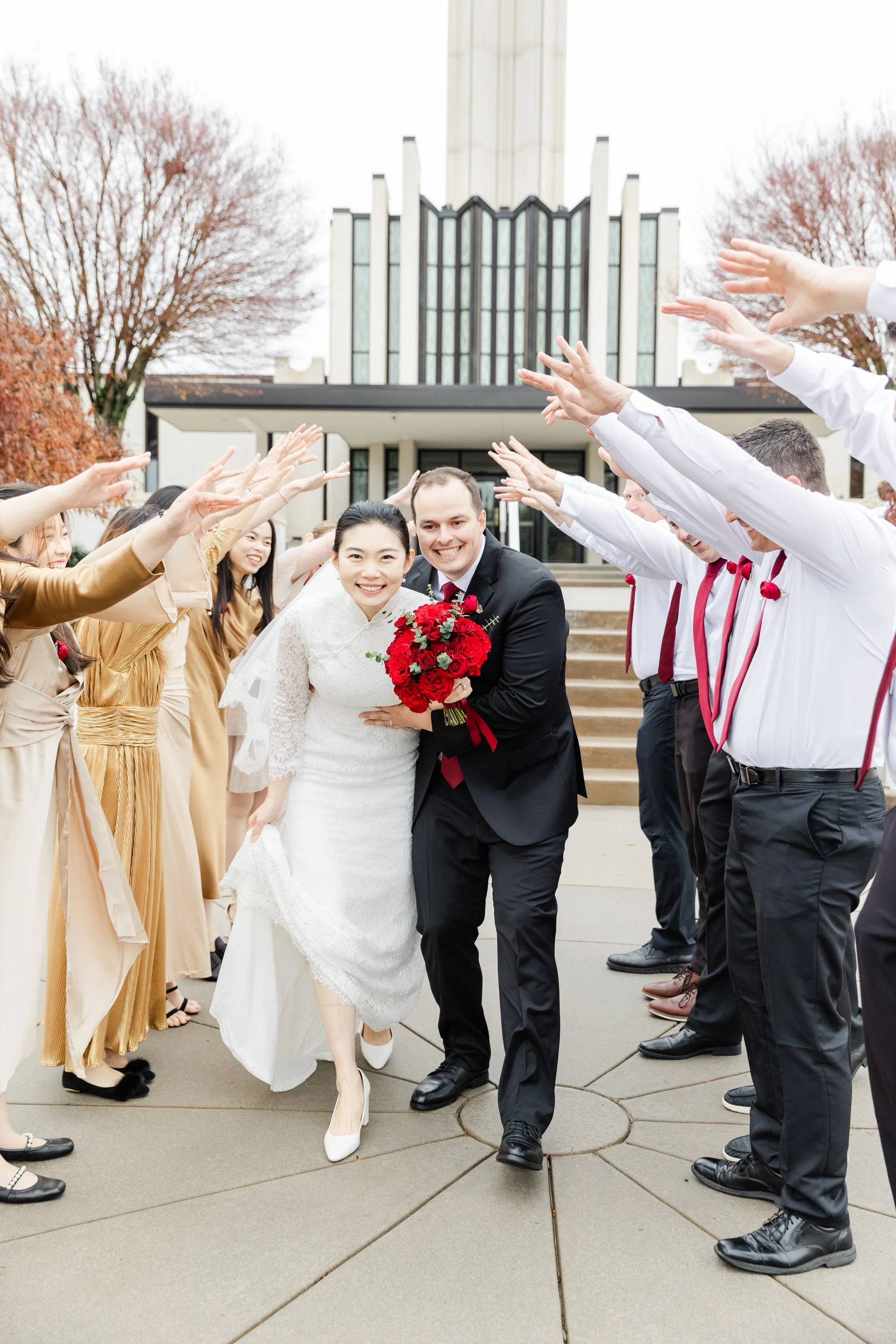 A newlywed couple walking through a line of friends and family during their wedding celebration outside a modern church, with everyone raising their hands in a celebratory gesture.