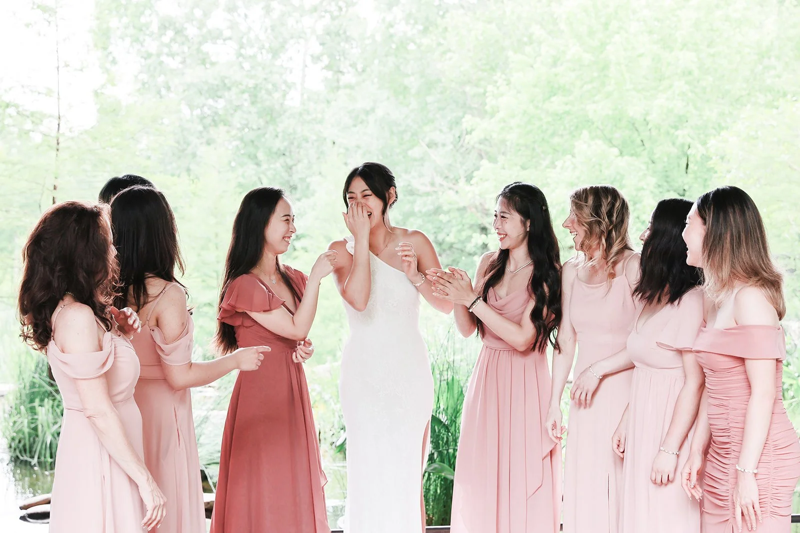 A bride in a white dress is surrounded by six women in pink dresses, all smiling and laughing, in an outdoor setting with green trees in the background.