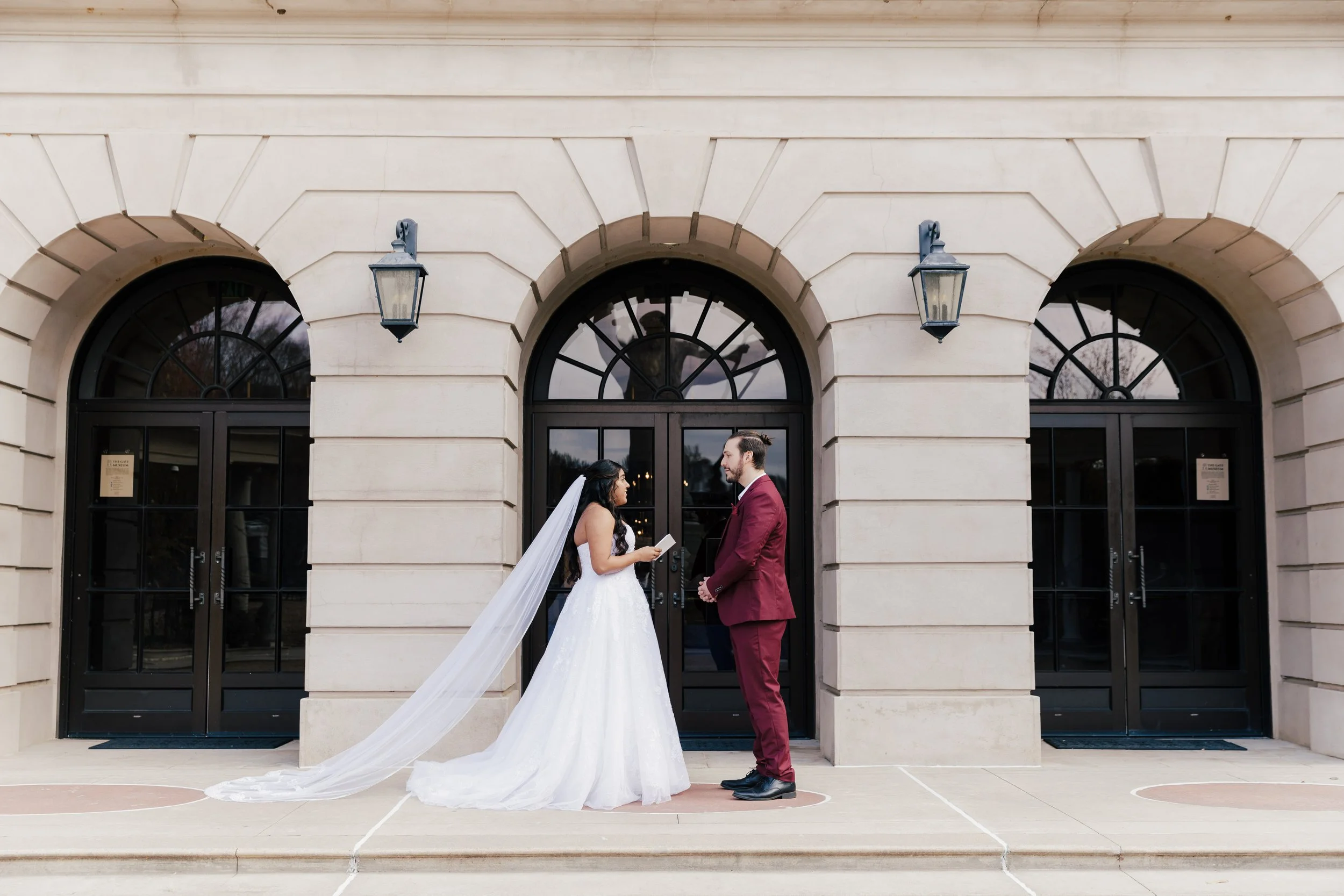 Bride and groom standing in front of a building, facing each other, during their wedding ceremony. The bride is wearing a white wedding dress with a long veil, and the groom is wearing a burgundy suit. The building has three arched doors with black f
