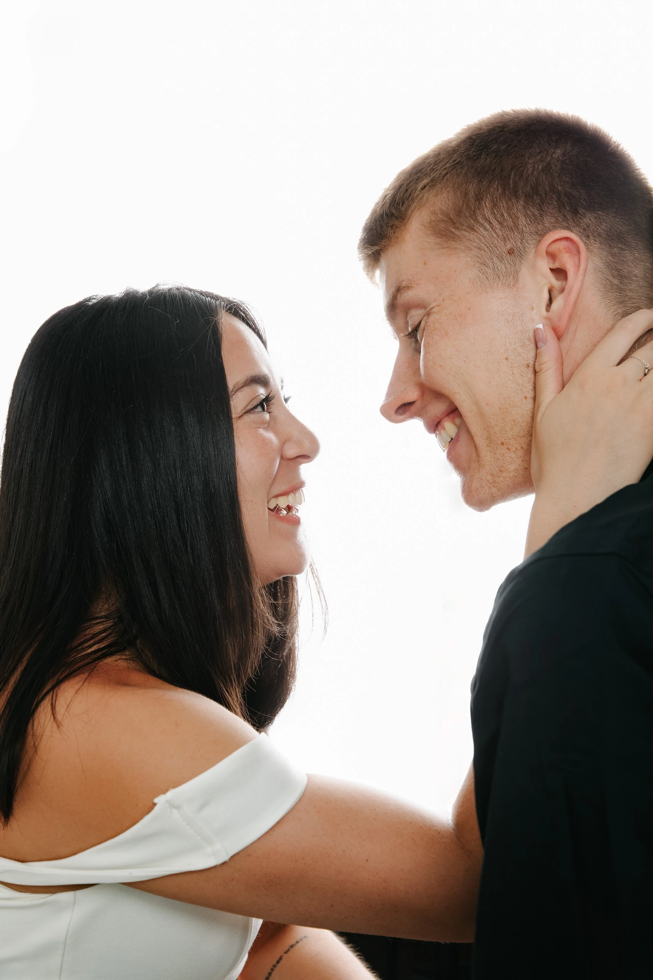 A smiling couple facing each other closely, with the woman touching the man's face, both appearing happy and affectionate.