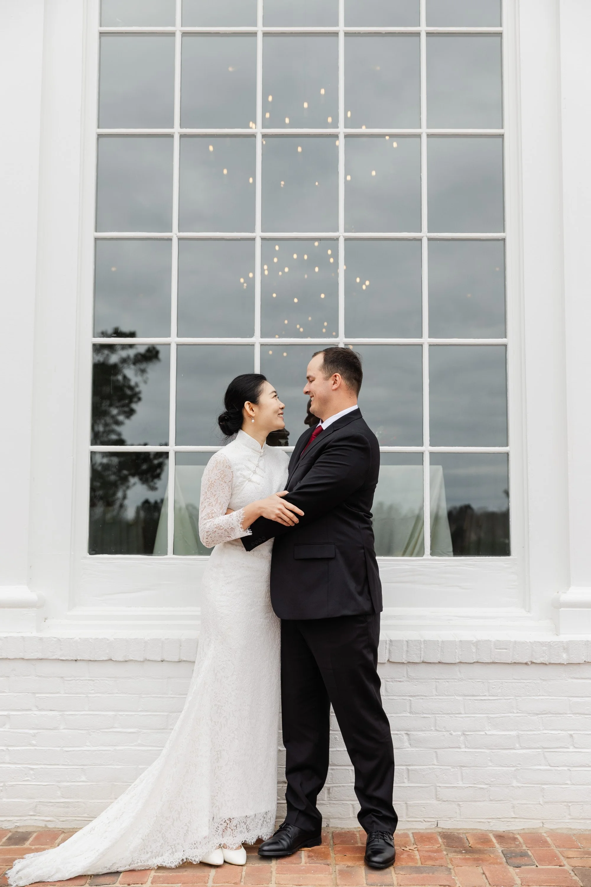 Bride and groom standing close and smiling at each other in front of a large window with a reflection of a chandelier and a cloudy sky.