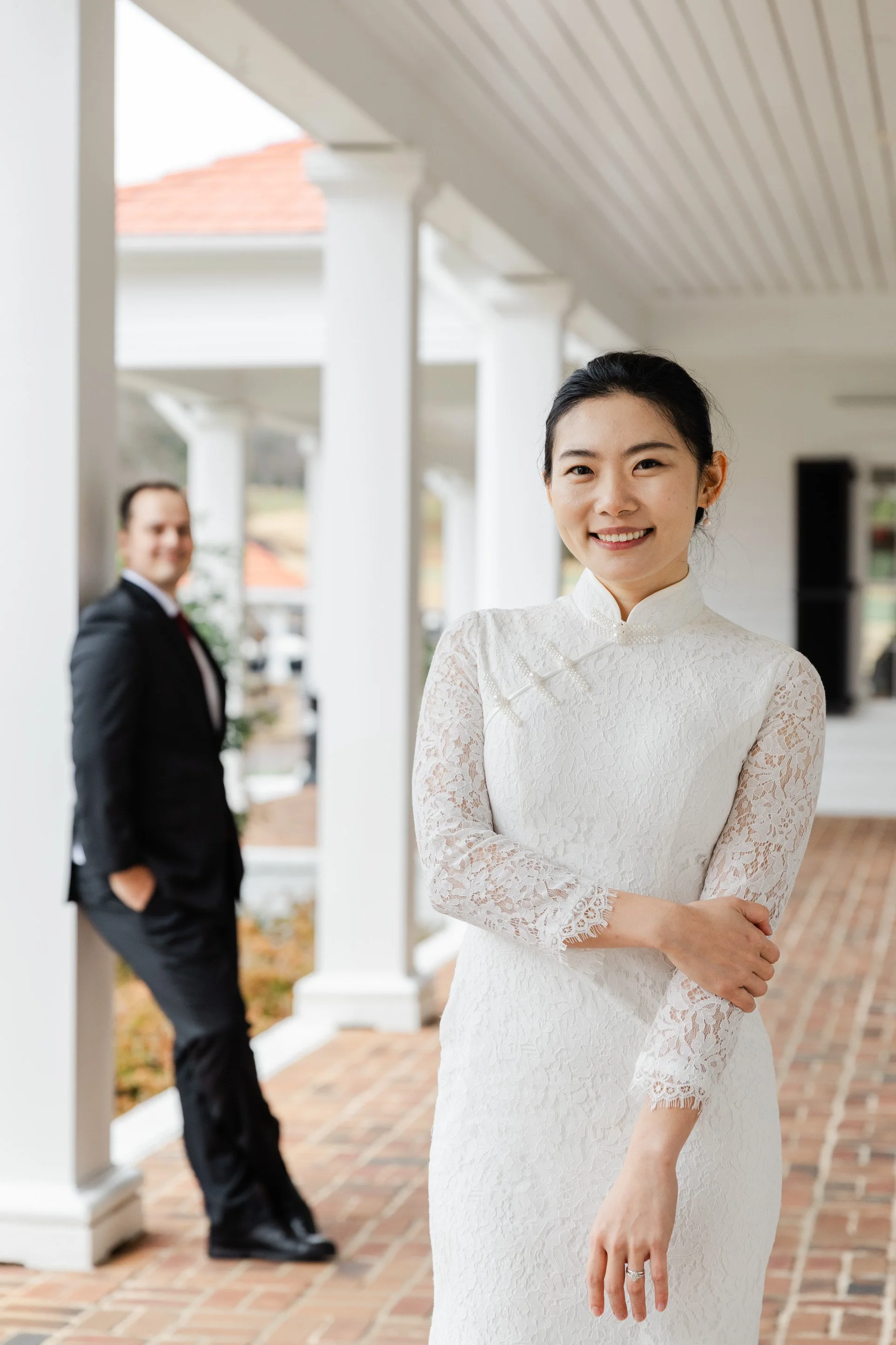 A smiling woman in a white lace dress standing outside on a porch, with a man in a suit blurred in the background.