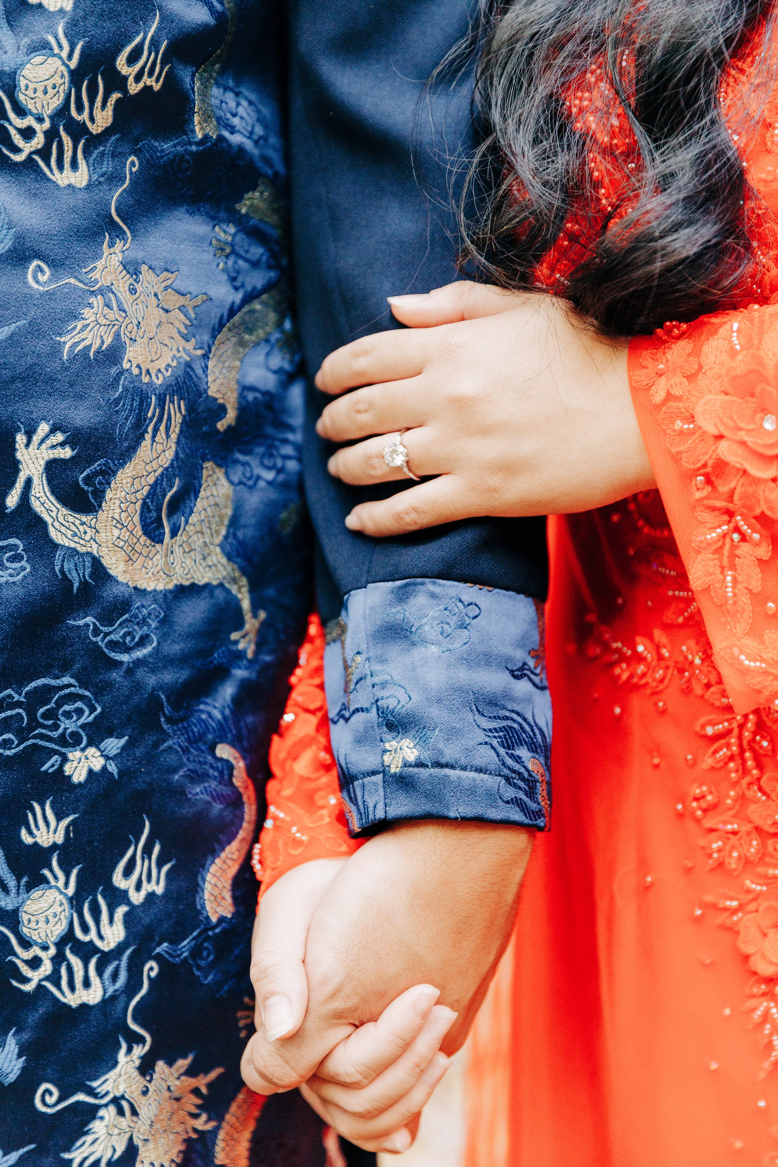 Close-up of a couple holding hands, with the woman's hand displaying an engagement ring, and both wearing traditional Asian attire, the man's outfit with gold dragon embroidery.