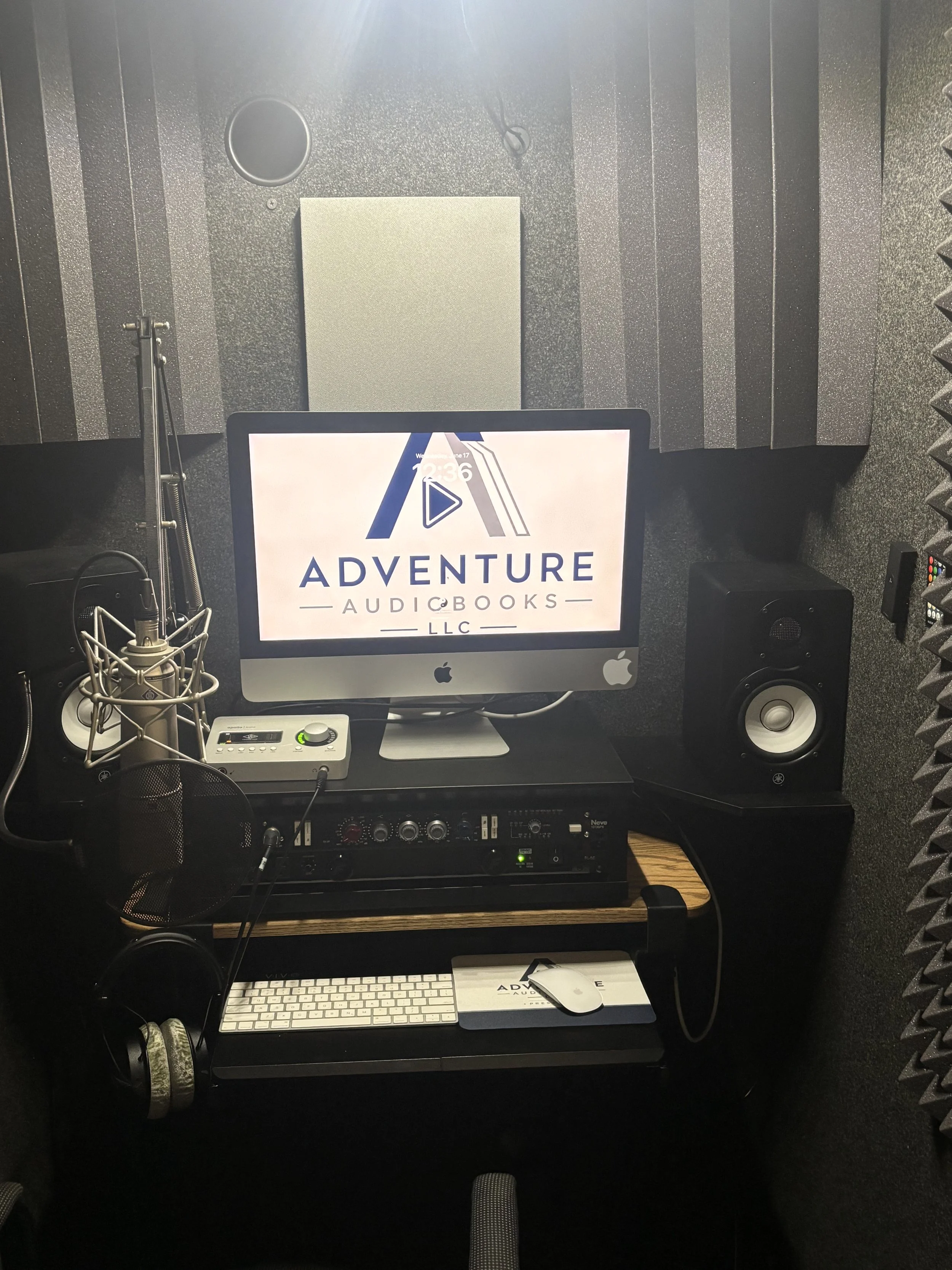 A recording studio setup with acoustic foam panels, a computer monitor displaying the 'Adventure Audiobooks LLC' logo, a microphone with a pop filter, a keyboard, a mouse, studio monitors, and various audio equipment.