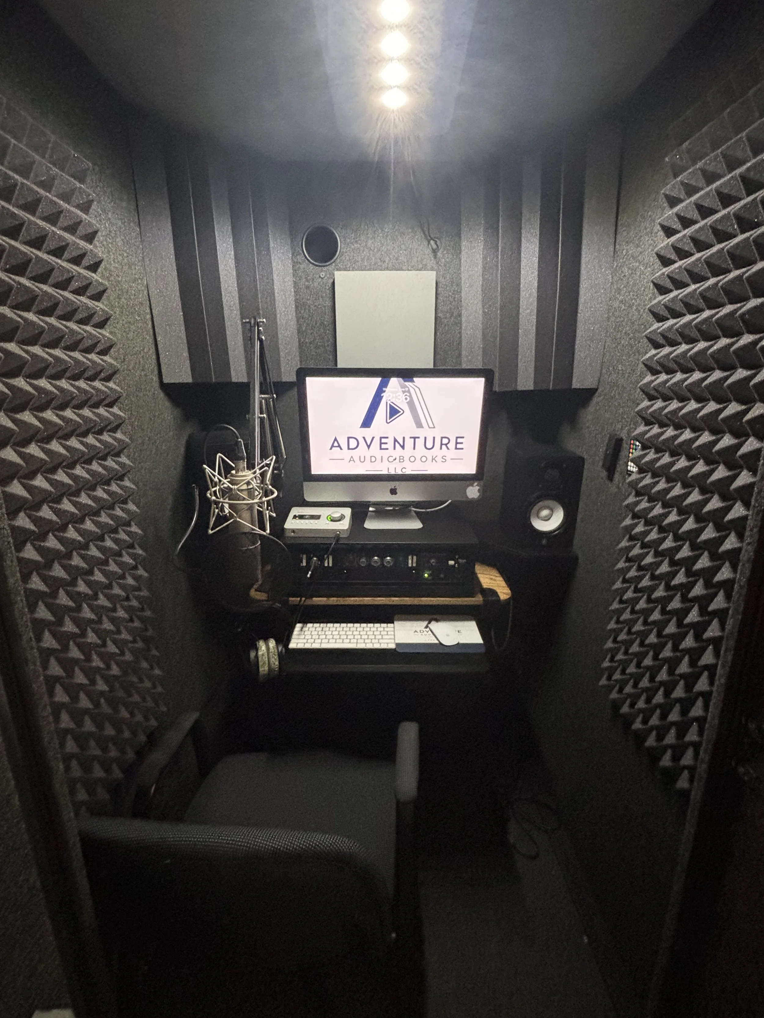 Soundproof recording booth with computer monitor, microphone, speaker, keyboard, and audio equipment.