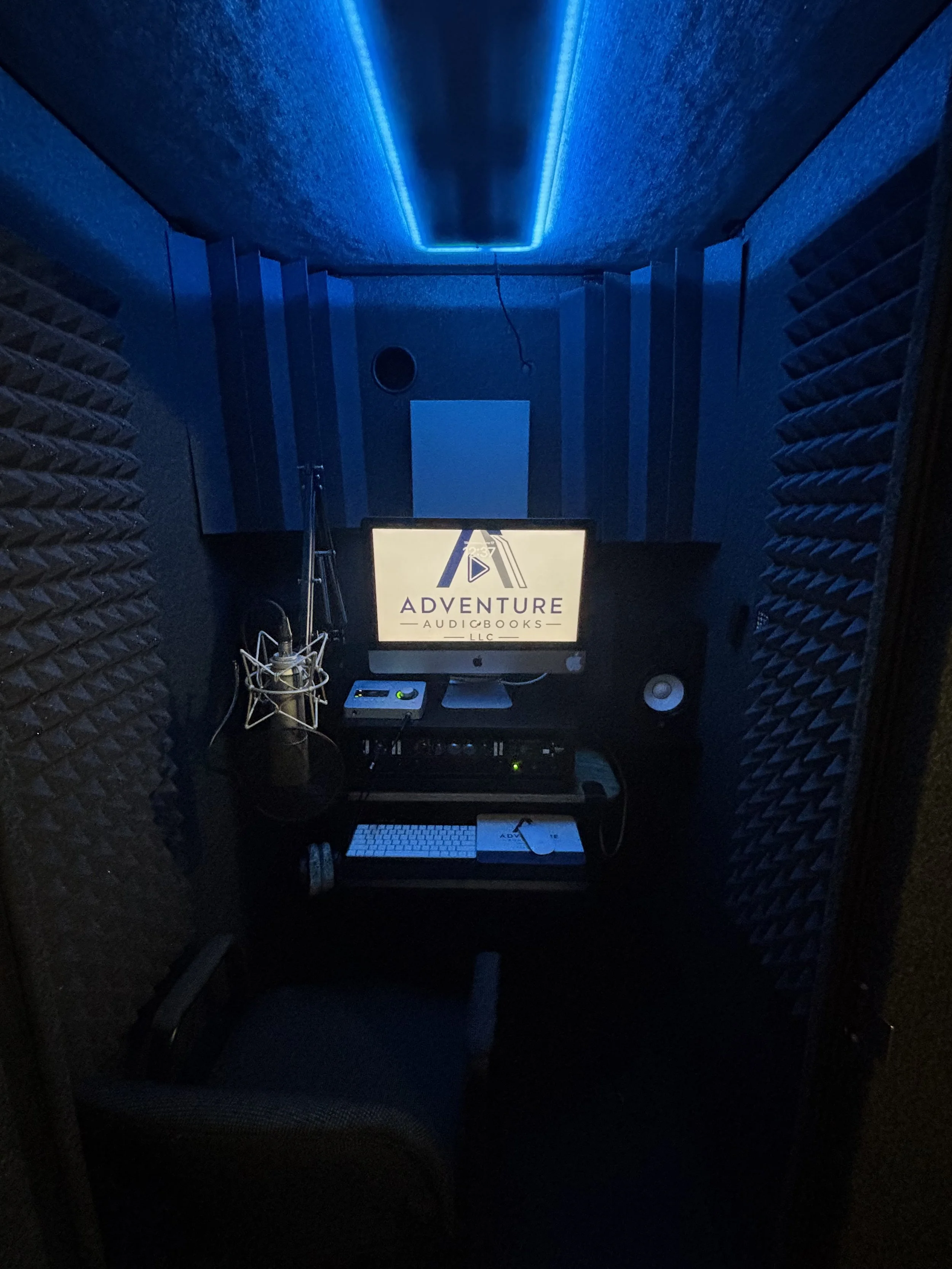 A professional recording studio with soundproofing foam walls, blue LED ceiling lights, a computer monitor displaying 'Adventure Audiobooks LLC,' a microphone, audio interface, and other audio equipment.