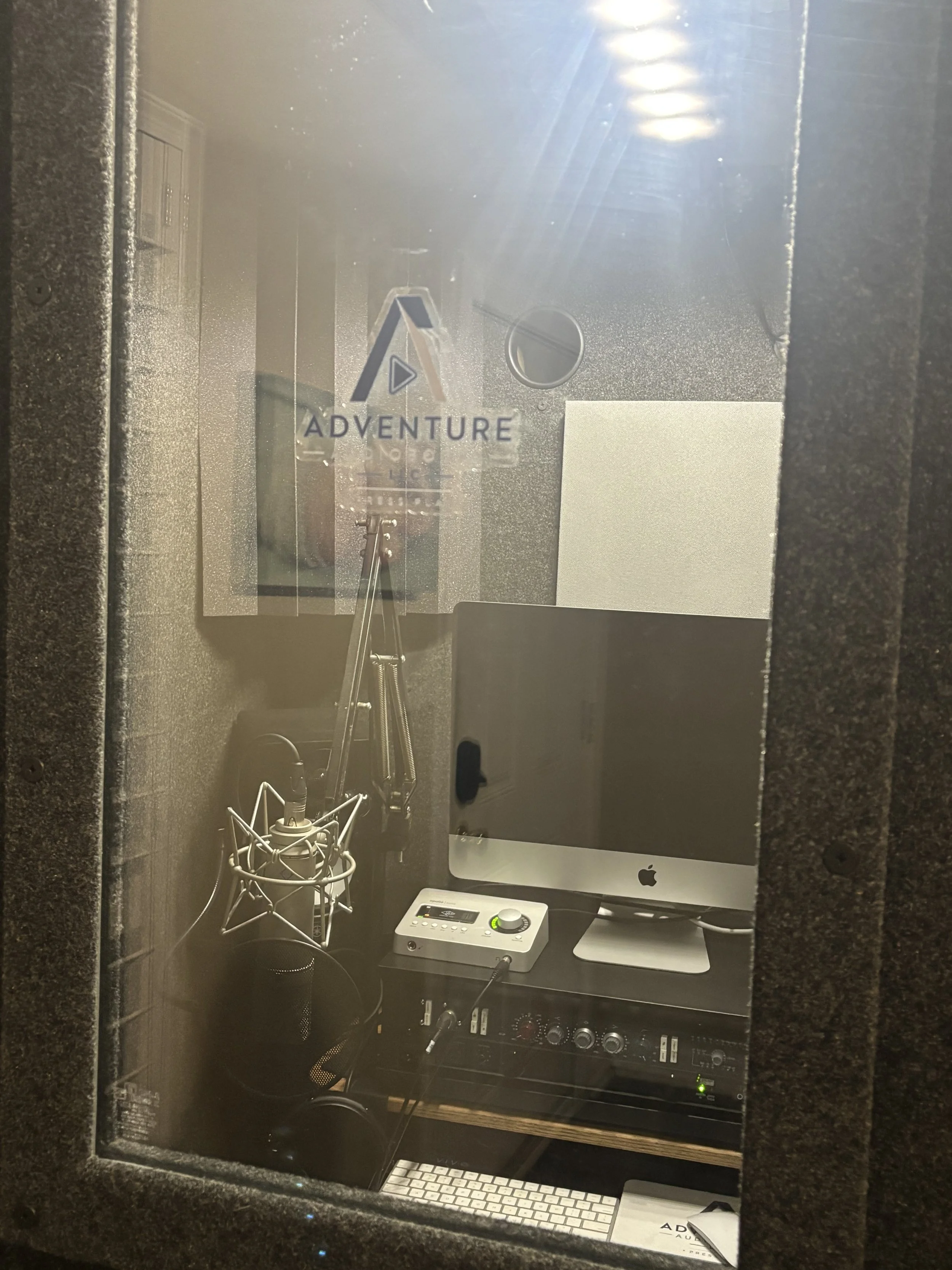 A recording studio booth with a microphone, computer, audio interface, and sound mixing equipment. The booth has a window with the logo 'Adventure' visible.