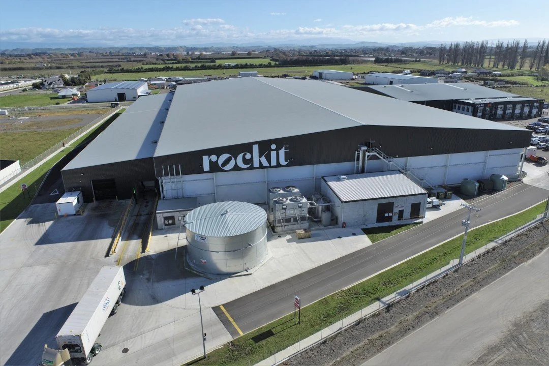 Rockit Apples Ironside - Industrial Engineering - Strata Group Engineers NZ | Aerial view of a large warehouse building with "rockit" logo on the side, surrounded by a paved area, machinery, and storage tanks, situated in a rural area.