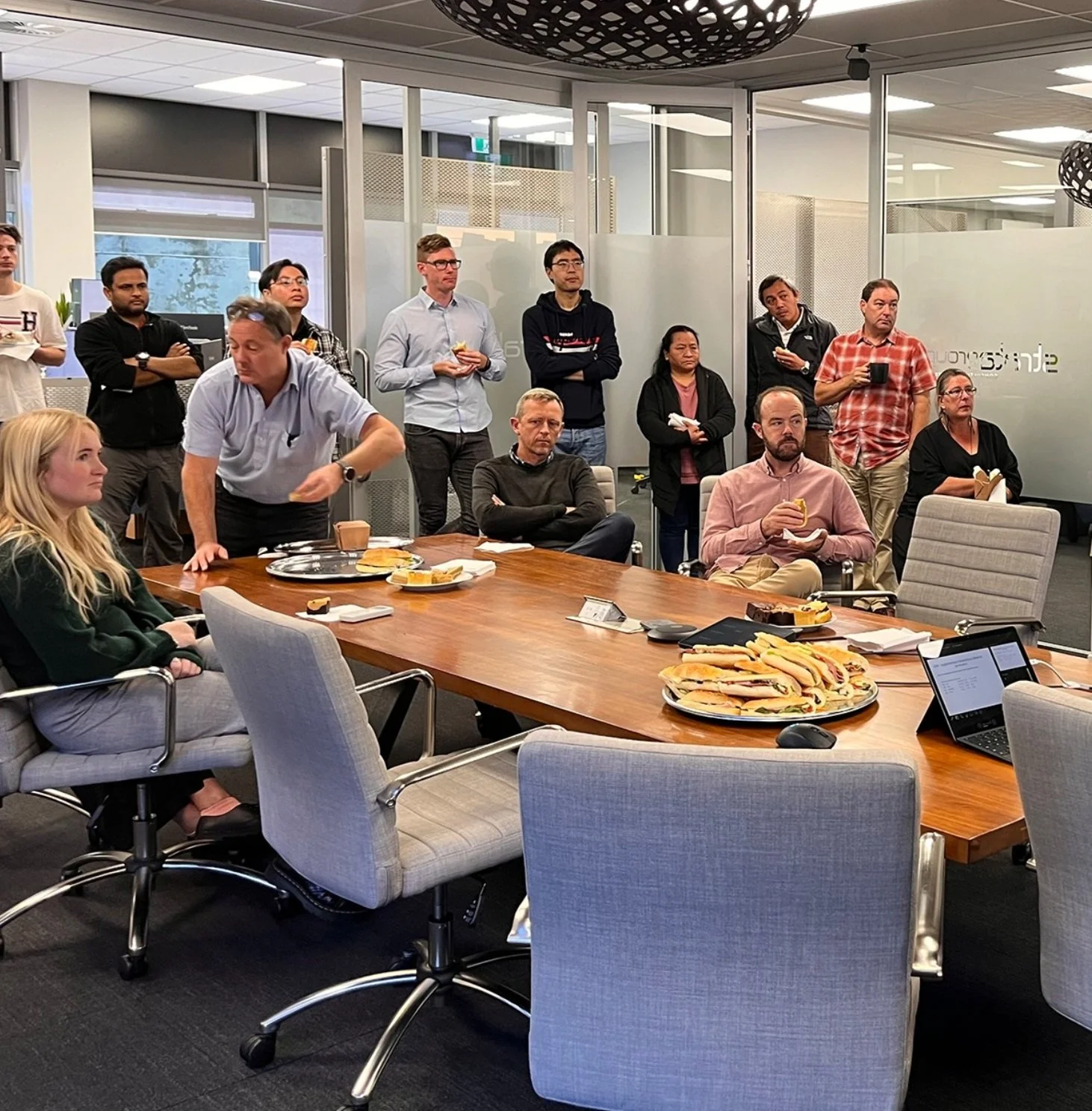 Our Team - Strata Group - New Zealand's Best Engineers | A group of people in an office meeting room, standing and sitting around a table with food and drinks. The room has glass walls with company branding.