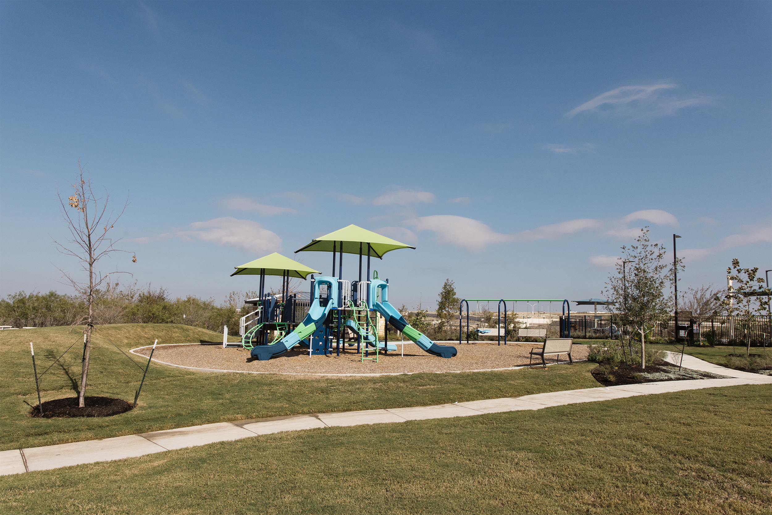 Playground with slides and swings surrounded by grass and trees under a blue sky.