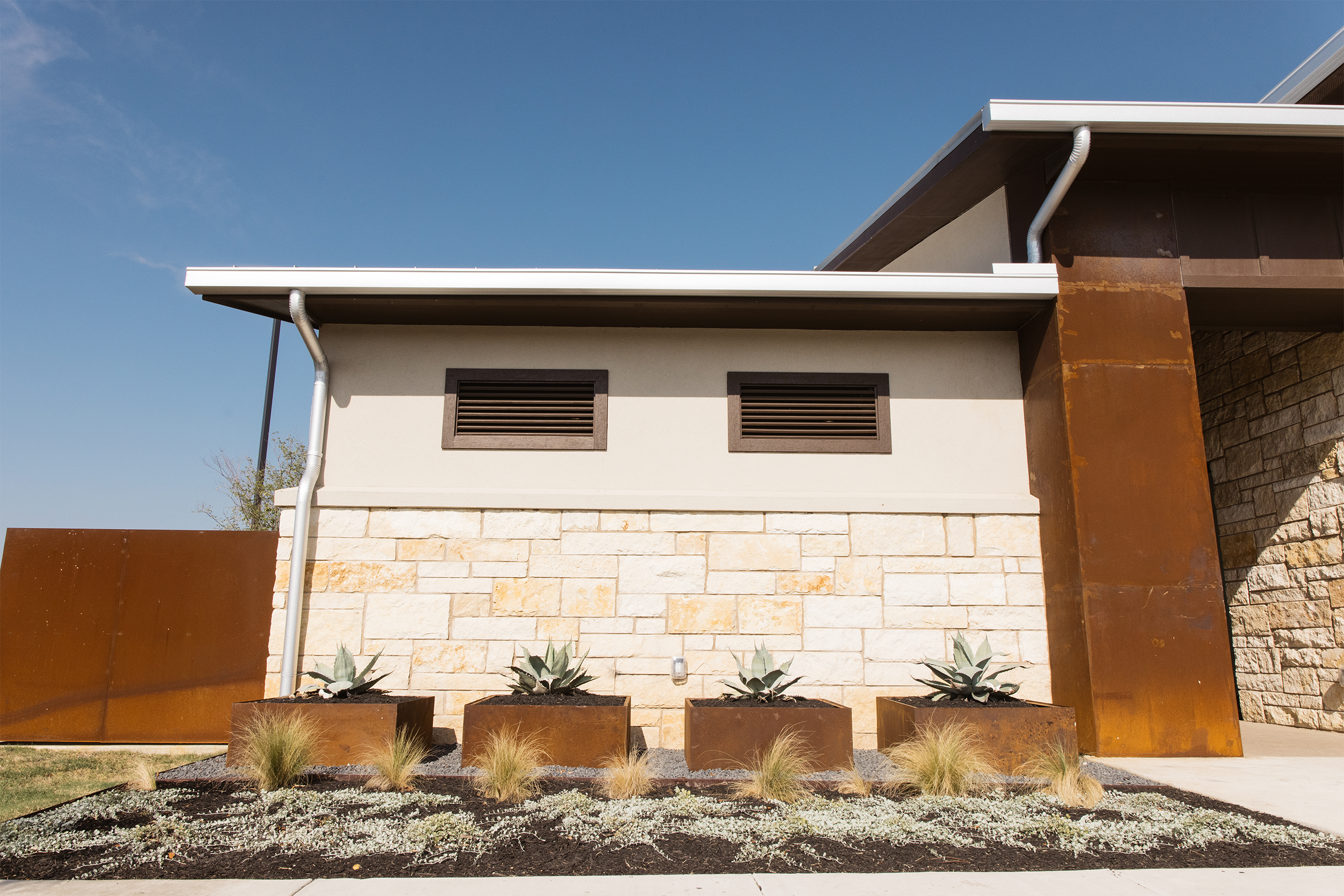 Modern building facade with stone and rust-colored metal paneling, featuring small windows with wooden slats. In the foreground, three large raised planters with succulent plants and surrounding landscaped area with grass and ground cover. Clear blue
