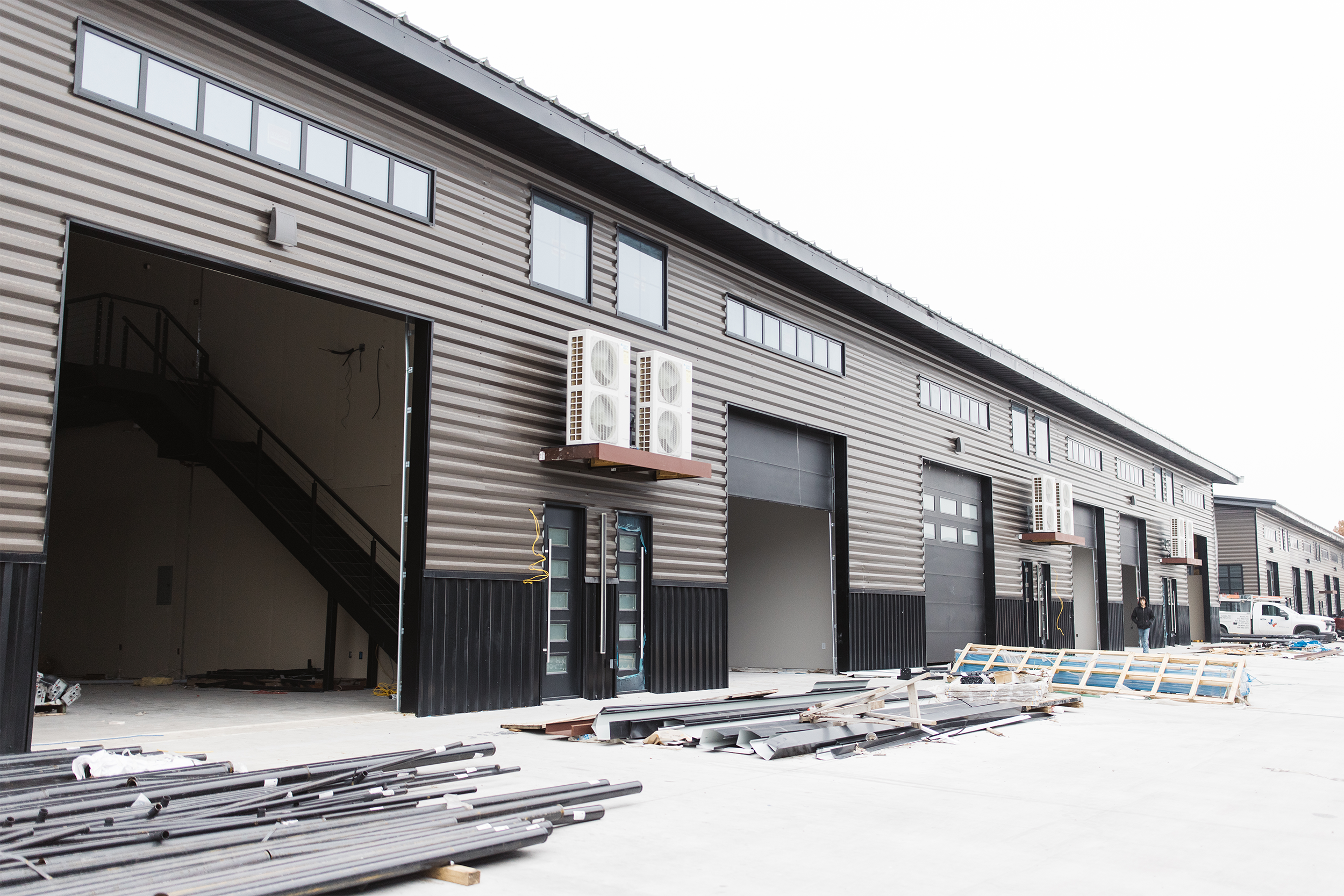 Outdoor view of a row of warehouse units under construction, featuring open garage doors and some installed HVAC units on a corrugated metal facade. Building materials are scattered on the ground.