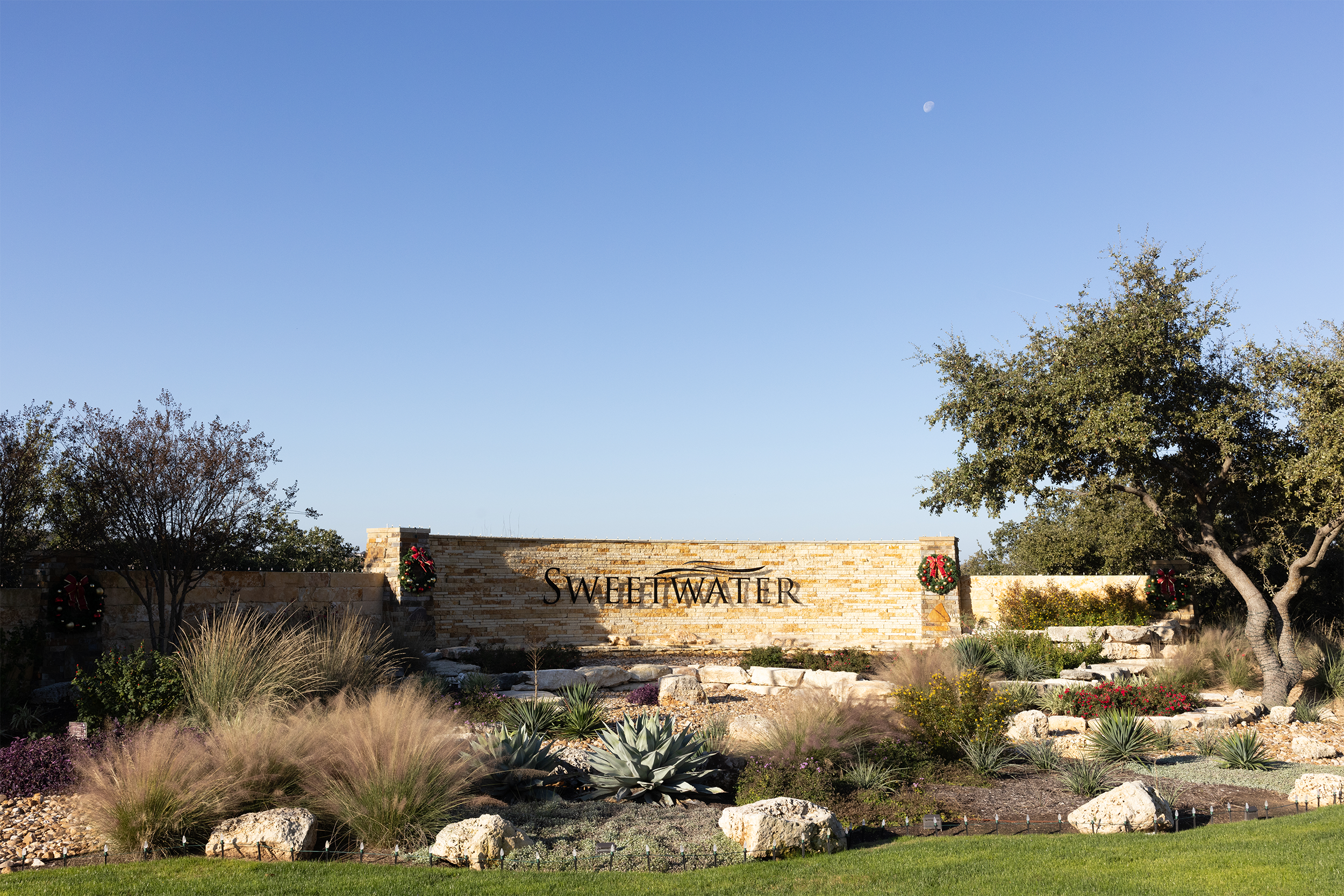 Entrance sign for Sweetwater community surrounded by landscaped garden featuring rocks, grasses, and trees, against a clear blue sky.