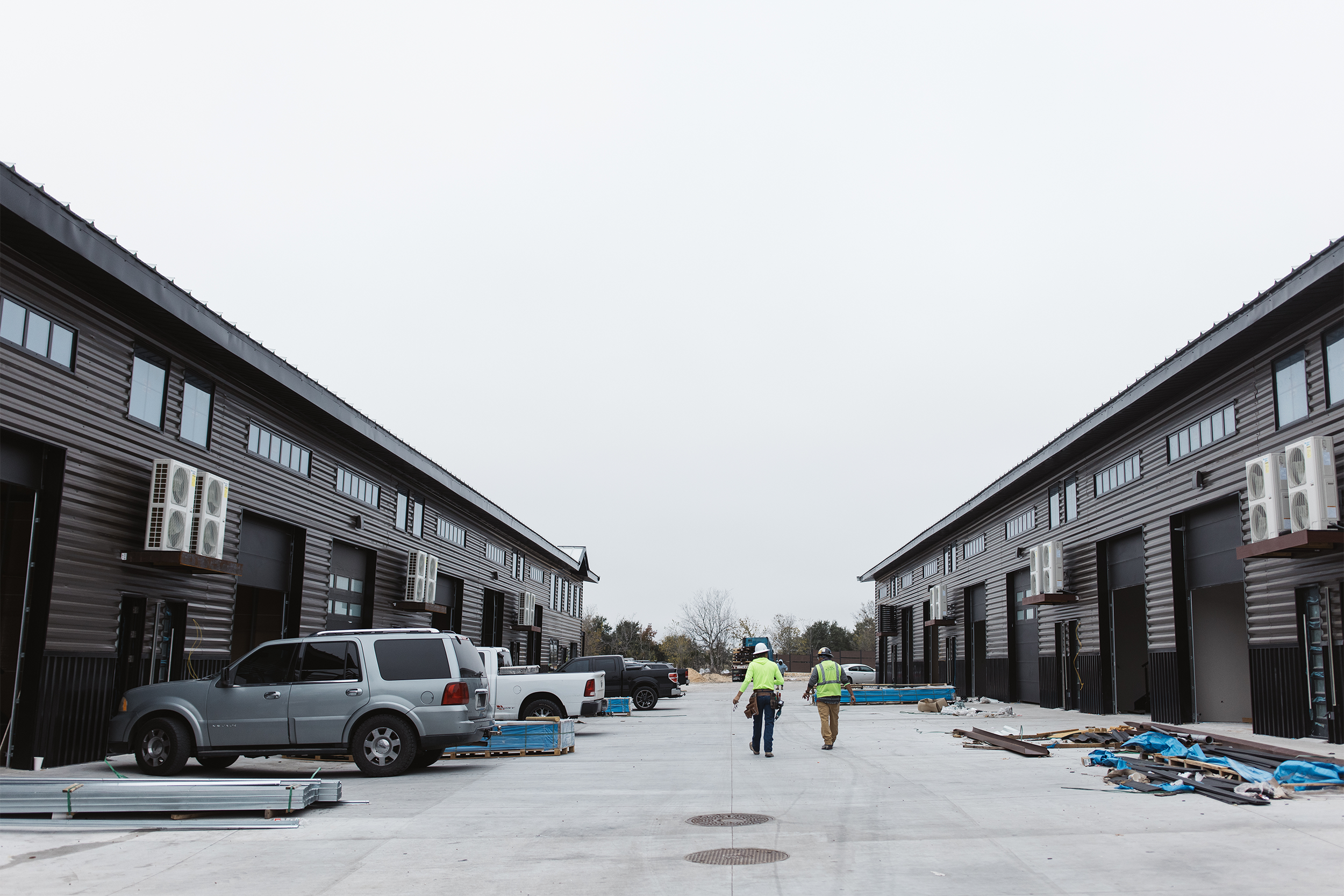 Two people wearing safety vests walking between two rows of industrial warehouse buildings with parked vehicles and construction materials around.