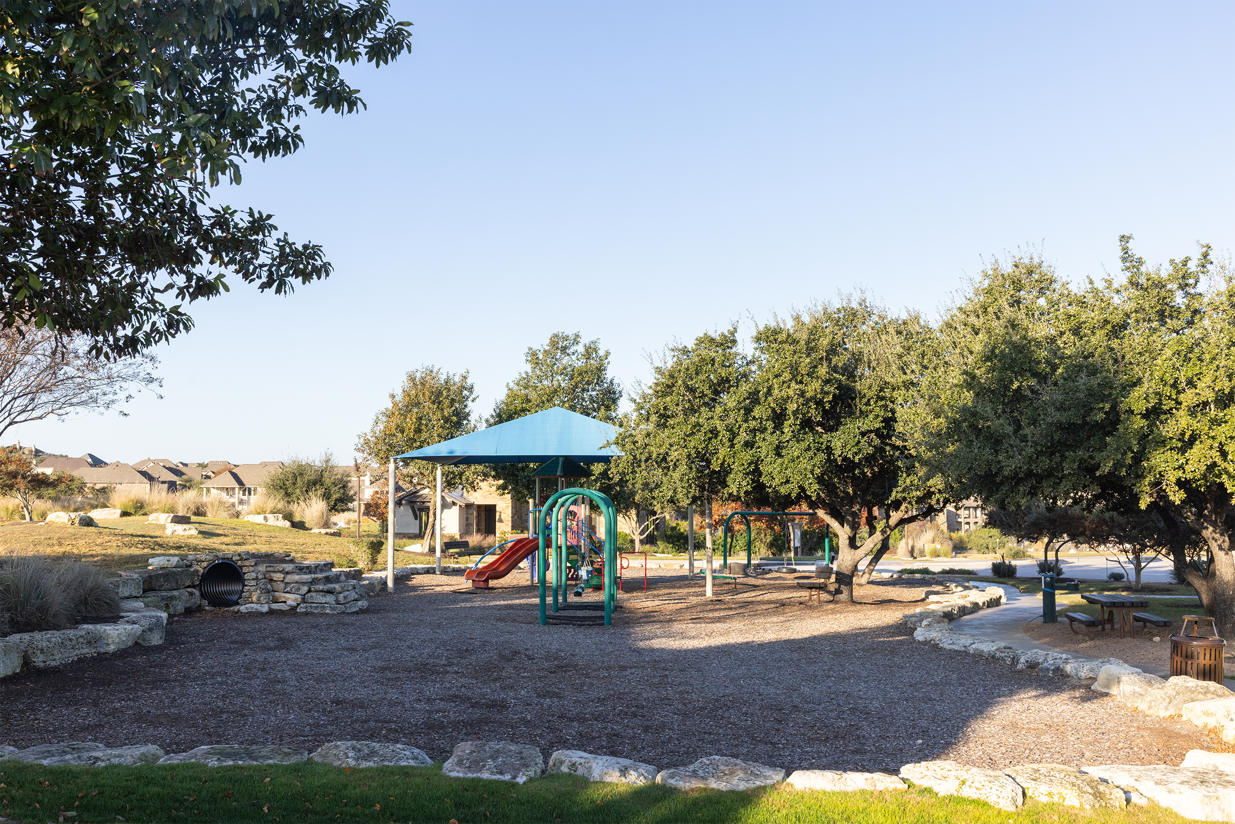Empty playground in a park with a blue shade canopy, surrounded by trees and residential houses in the background.