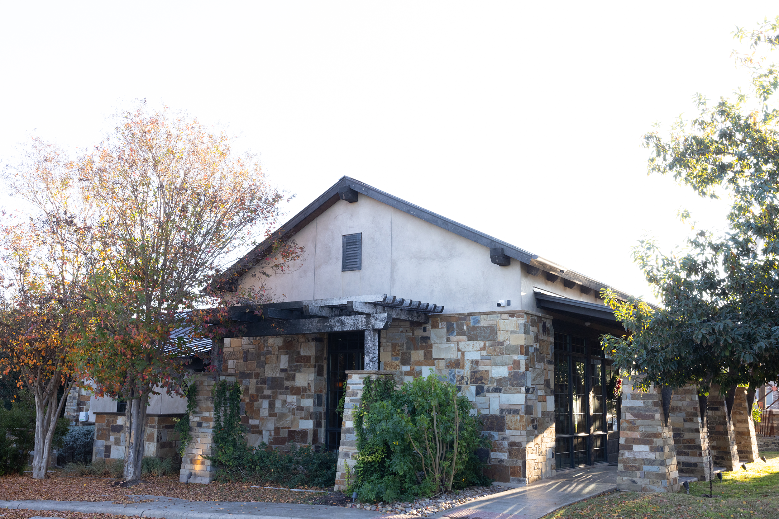 A small building with stone walls, a wooden pergola, and surrounding trees with autumn foliage.