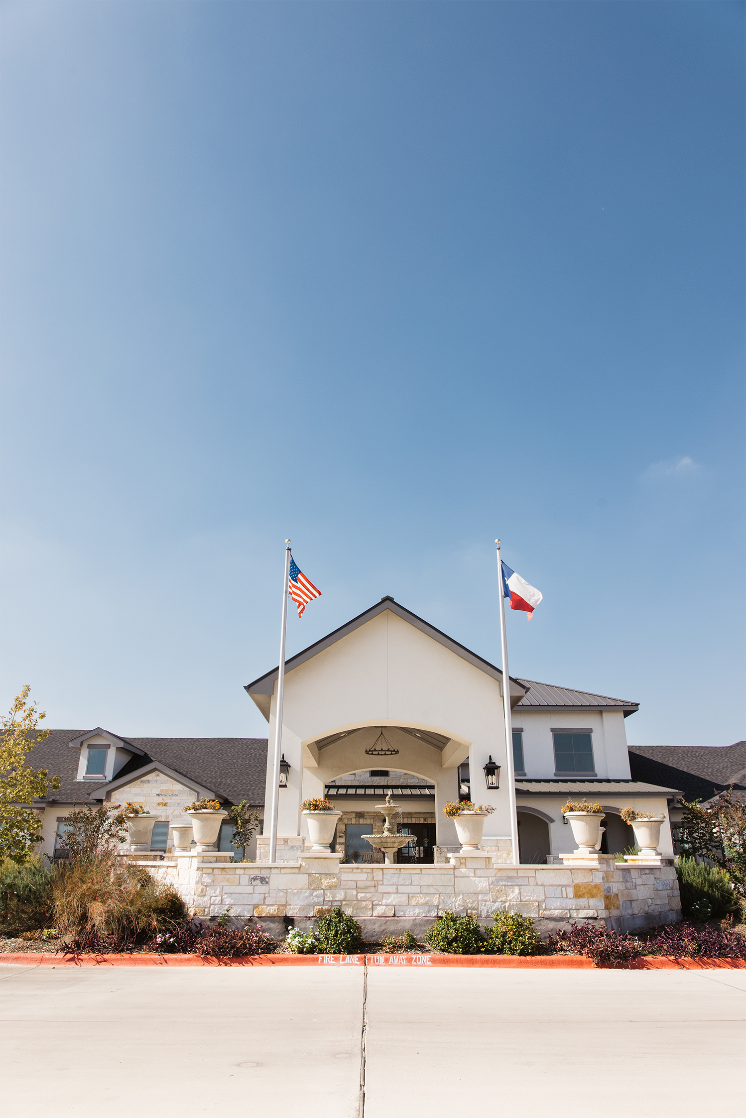 Facade of a house with two flagpoles displaying the American flag and another flag. The house has a stone wall, columns with flower pots, and a central fountain. Bright blue sky overhead.