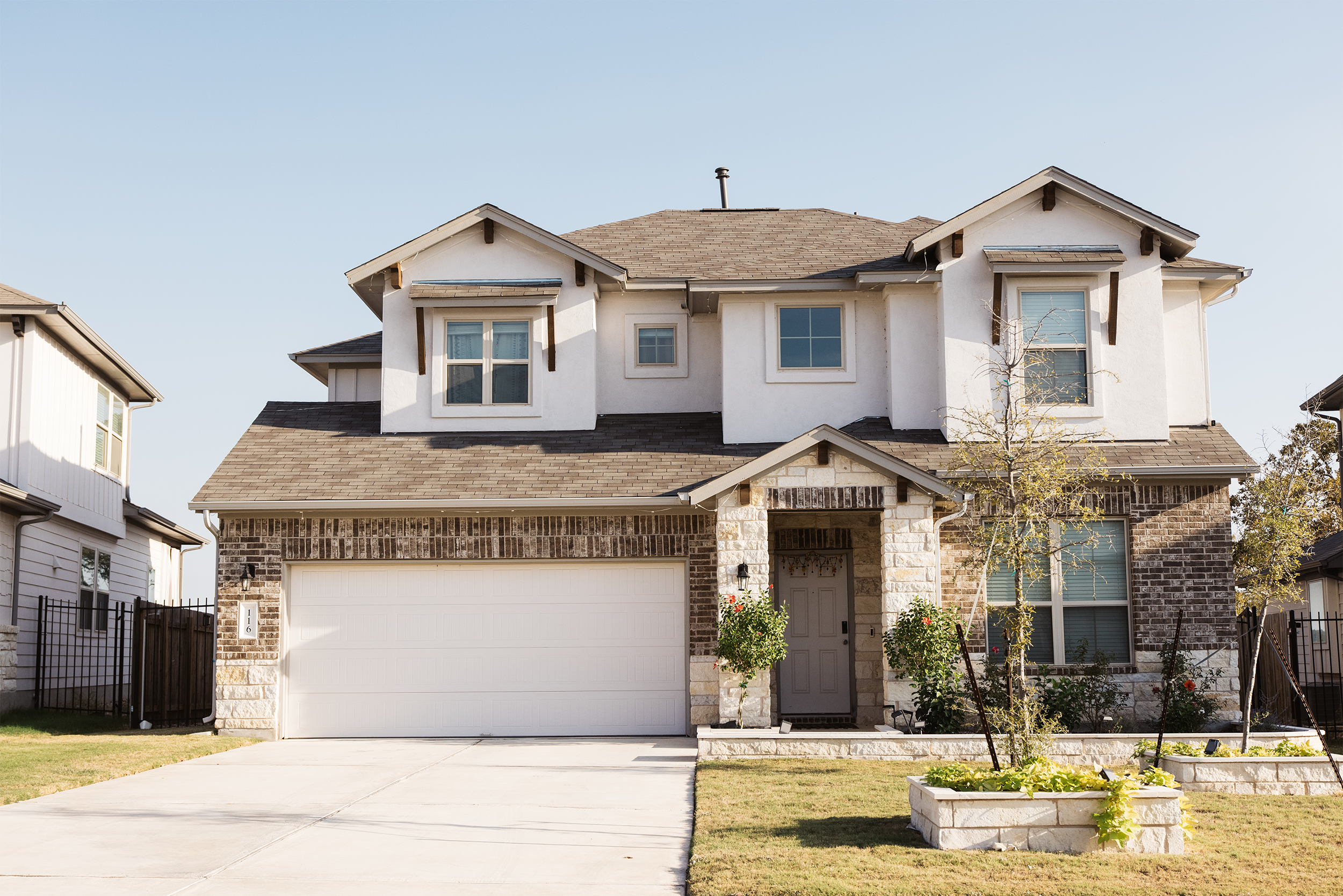 Two-story suburban house with a tan facade, gabled roof, and attached garage. The front yard has small trees and planter boxes.