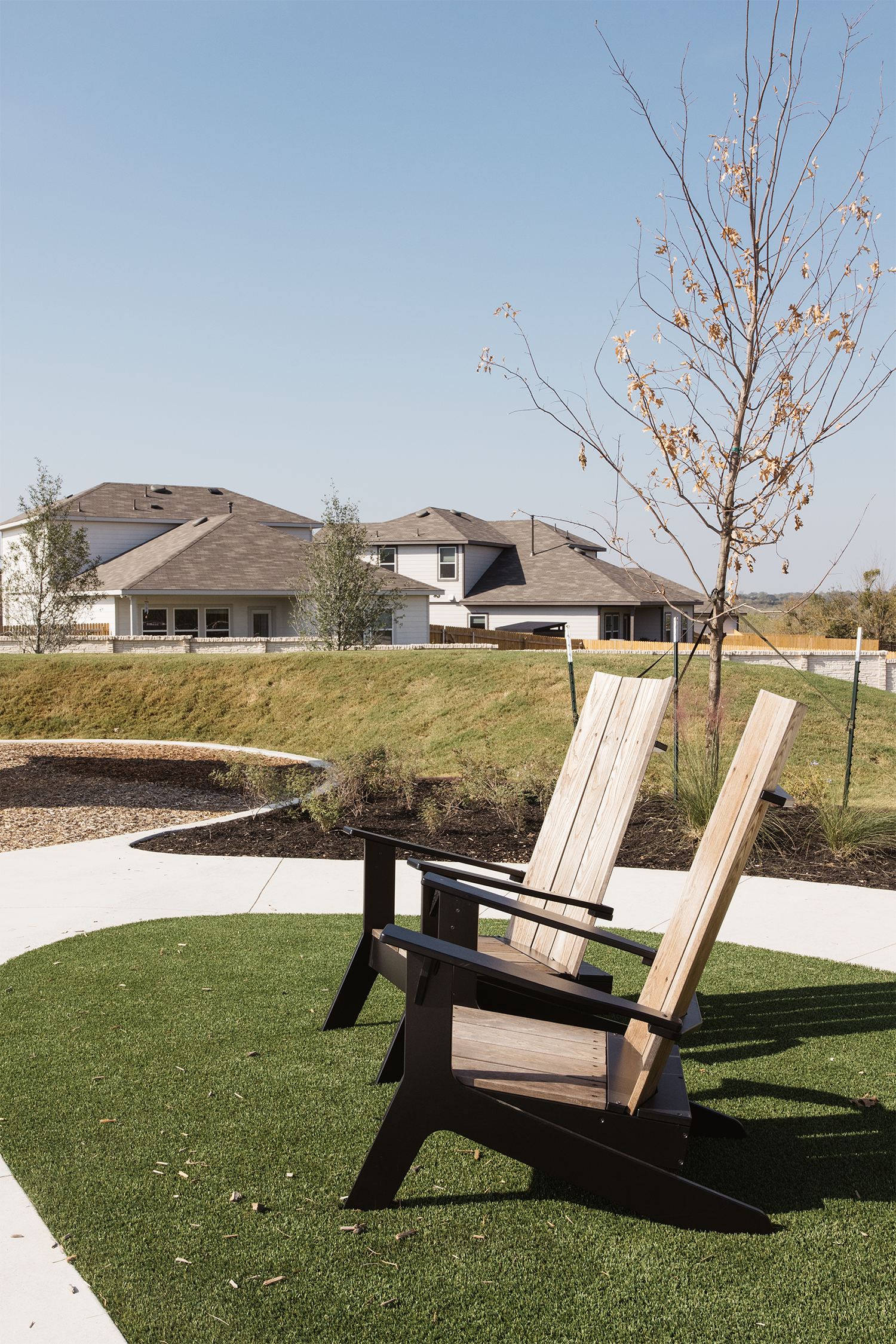 Two wooden chairs on grass with houses and trees in the background, under a clear blue sky.