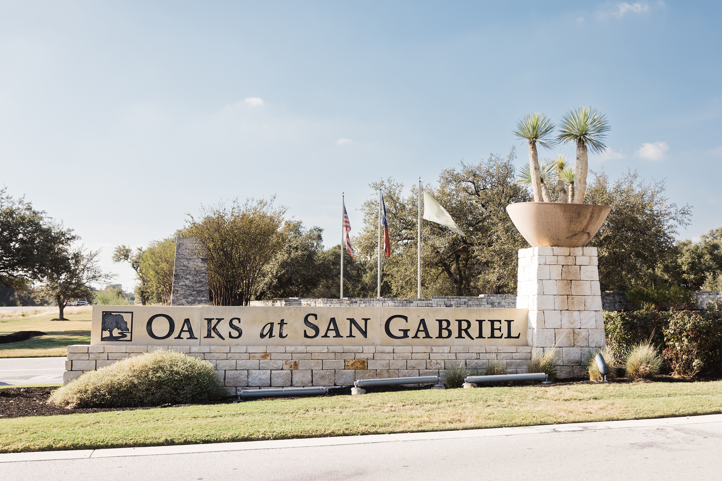 Entrance sign for Oaks at San Gabriel with stone pillars and landscaping, featuring three flags in the background.