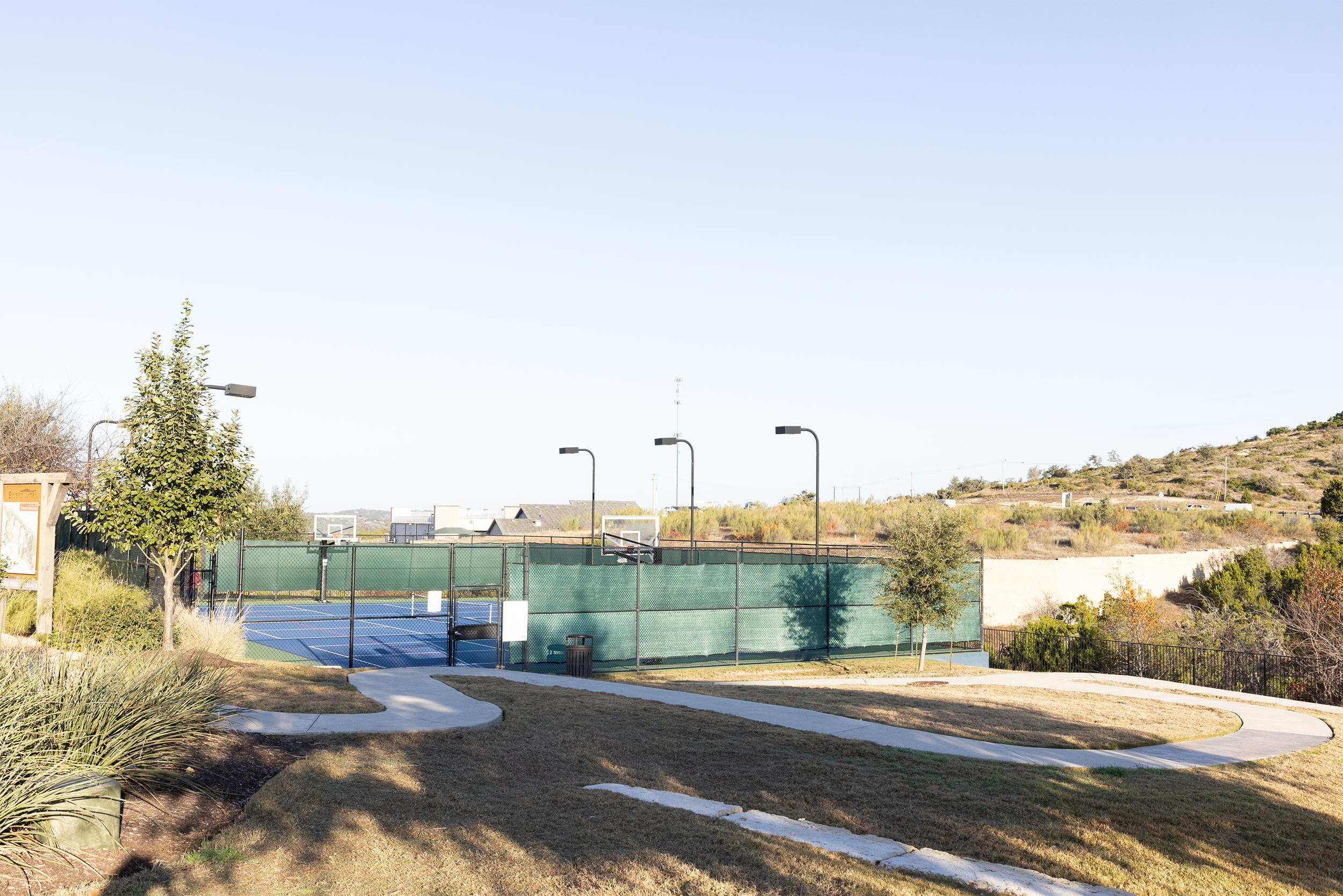 Outdoor sports court with basketball hoops and fencing, surrounded by greenery and a walking path under a clear sky.