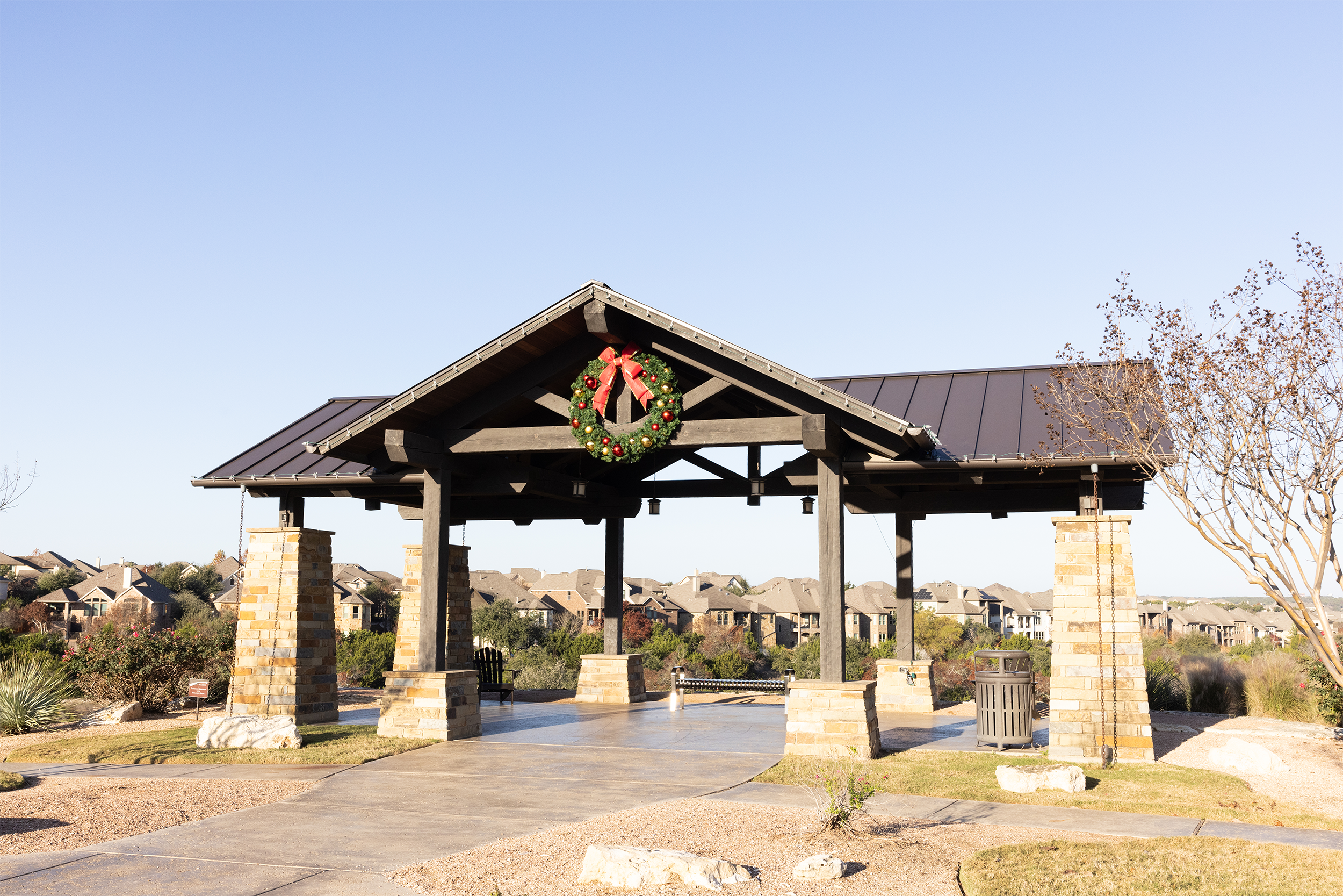 Outdoor pavilion with stone pillars and metal roof, featuring a Christmas wreath with a red bow on the front. Suburban houses and trees in the background.