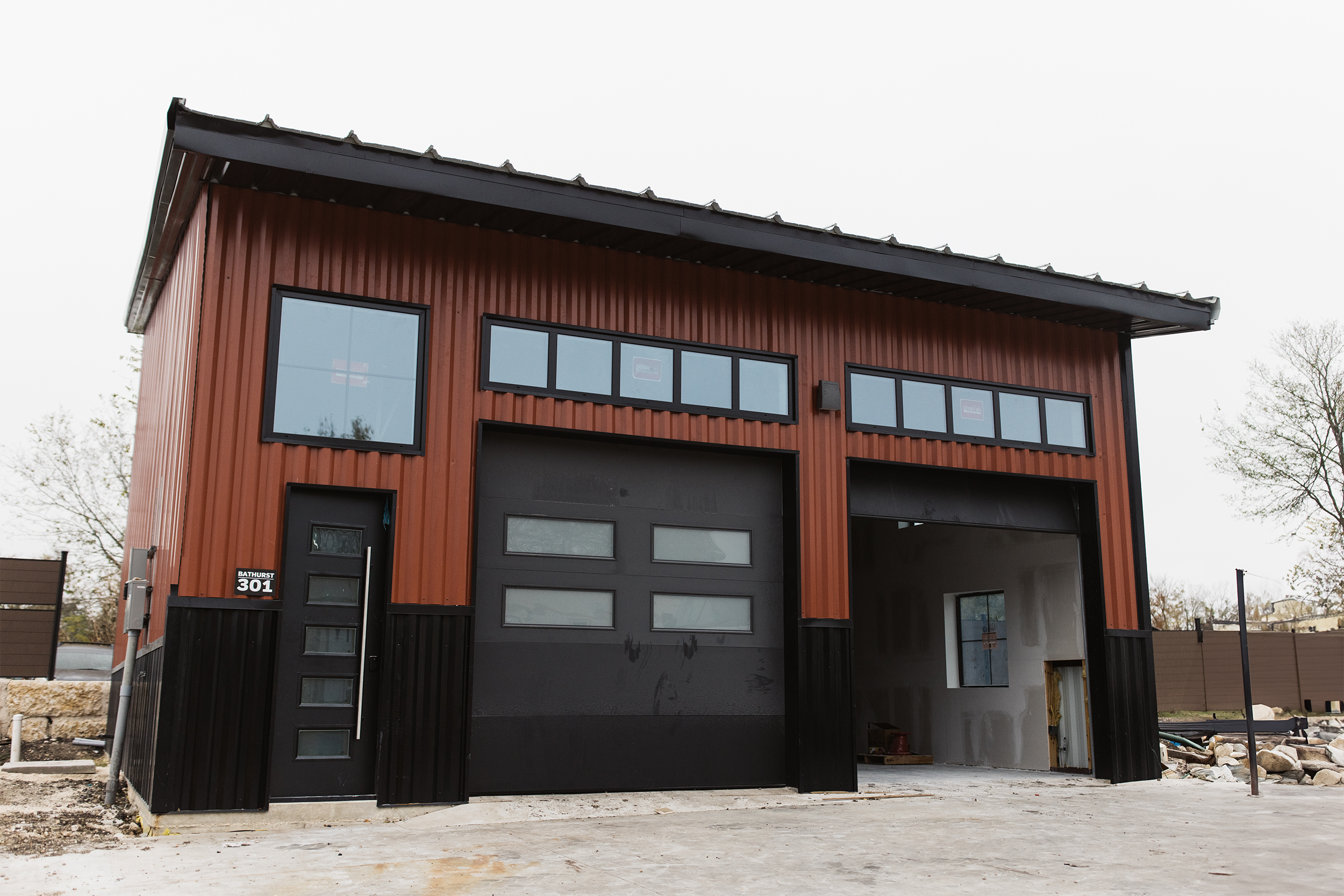Modern industrial-style garage with red and black metal siding, two large garage doors, and small upper windows. Located on a concrete lot with partially visible construction materials inside.