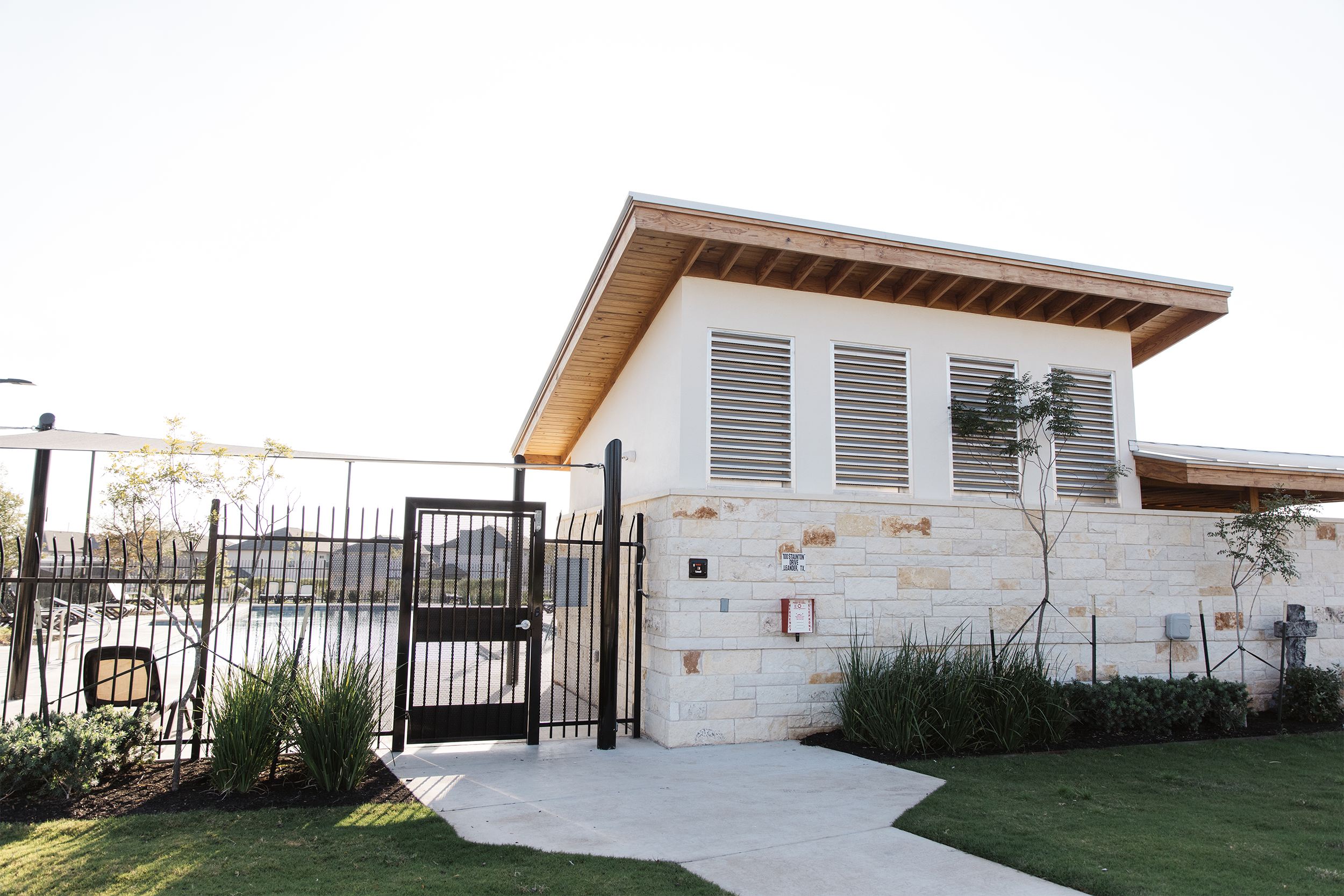 Single-story building with a gable roof, stone facade, and large metal gate in front, surrounded by landscaped greenery and a sidewalk.