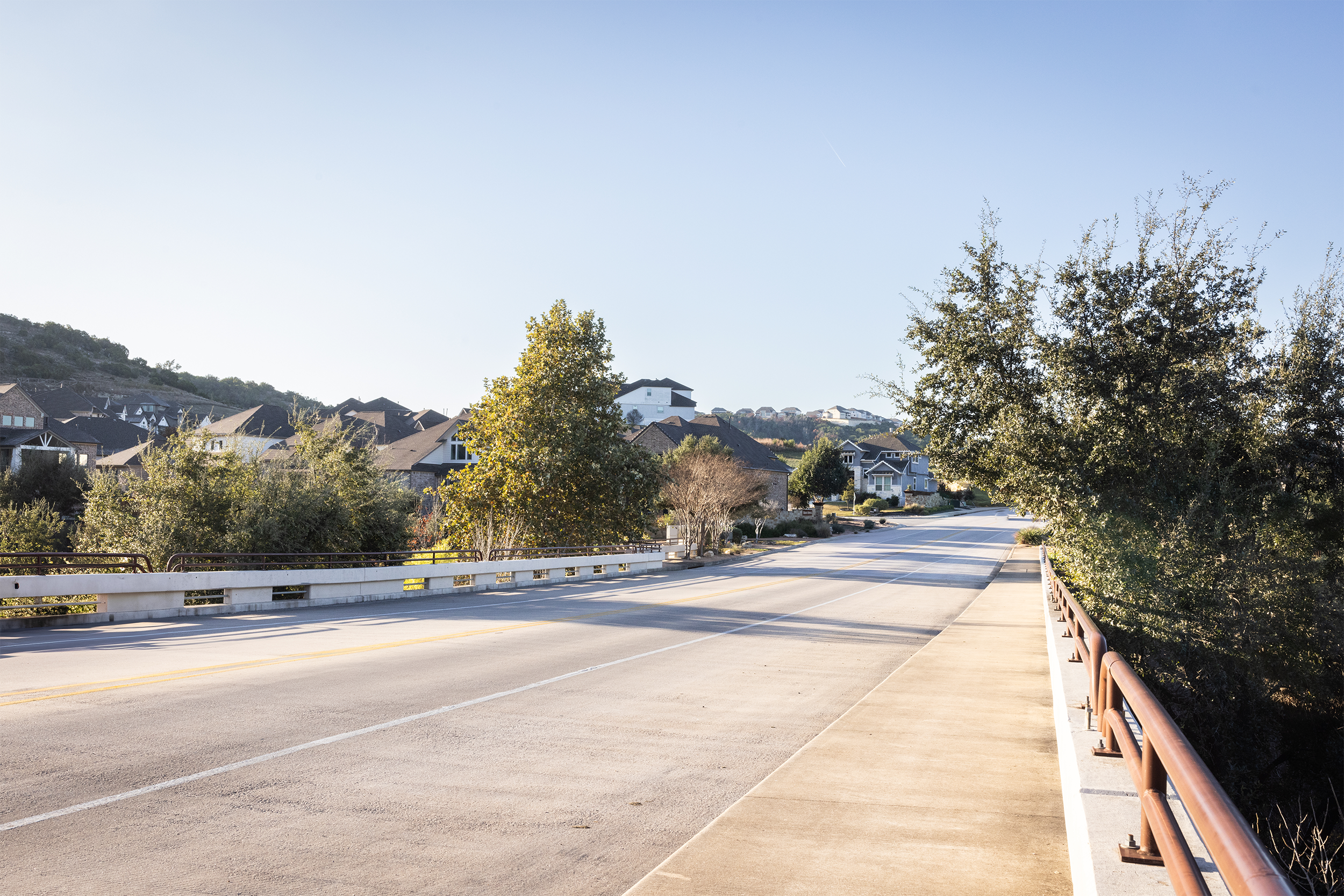 Residential neighborhood street view with houses, trees, and a bridge on a clear day.